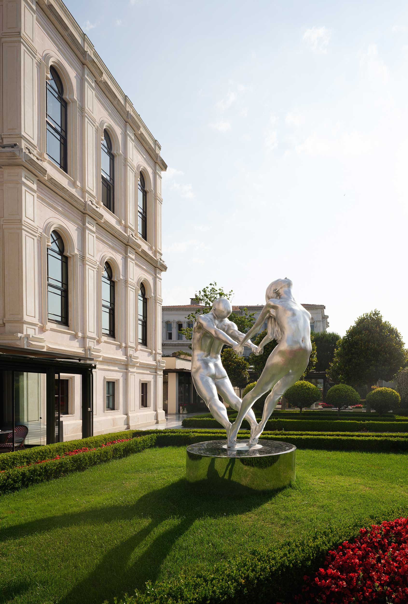 A modern sculpture of two intertwined figures holding hands, situated on a round black base in a landscaped garden with trimmed hedges and trees, with a historic building in the background on a sunny day at the Four Seasons Istanbul at the Bosphorus.