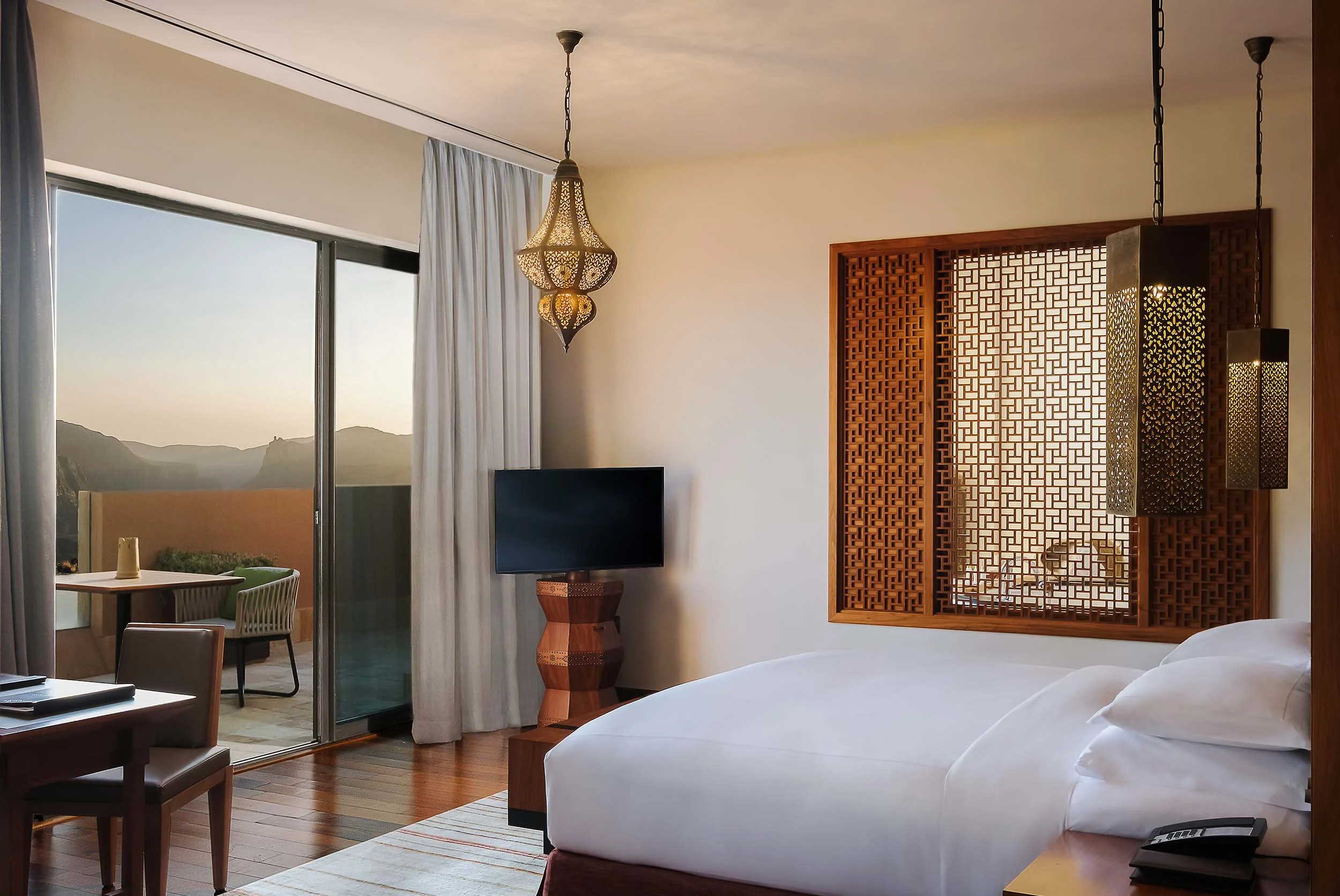 A hotel room with a large bed, a flat-screen TV on a wooden stand, and a sliding glass door leading to a balcony with outdoor seating and a mountain view.