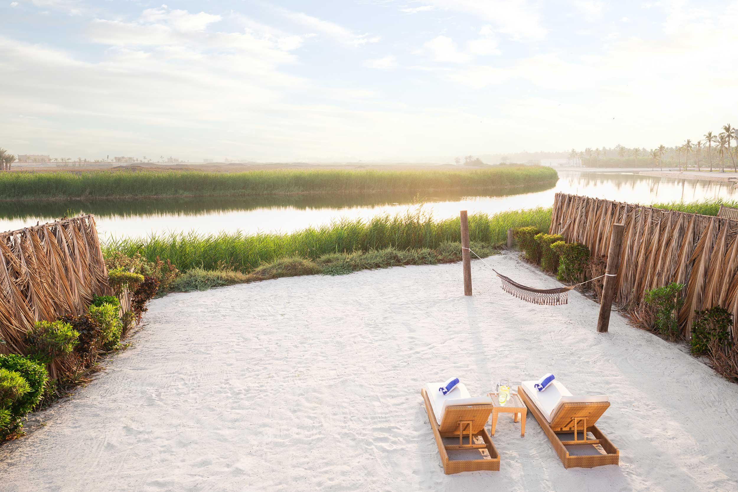 Beach scene with two lounge chairs, small table with drinks, hammock, sandy ground, and reed fences, overlooking calm water and green marshland with trees in the distance.