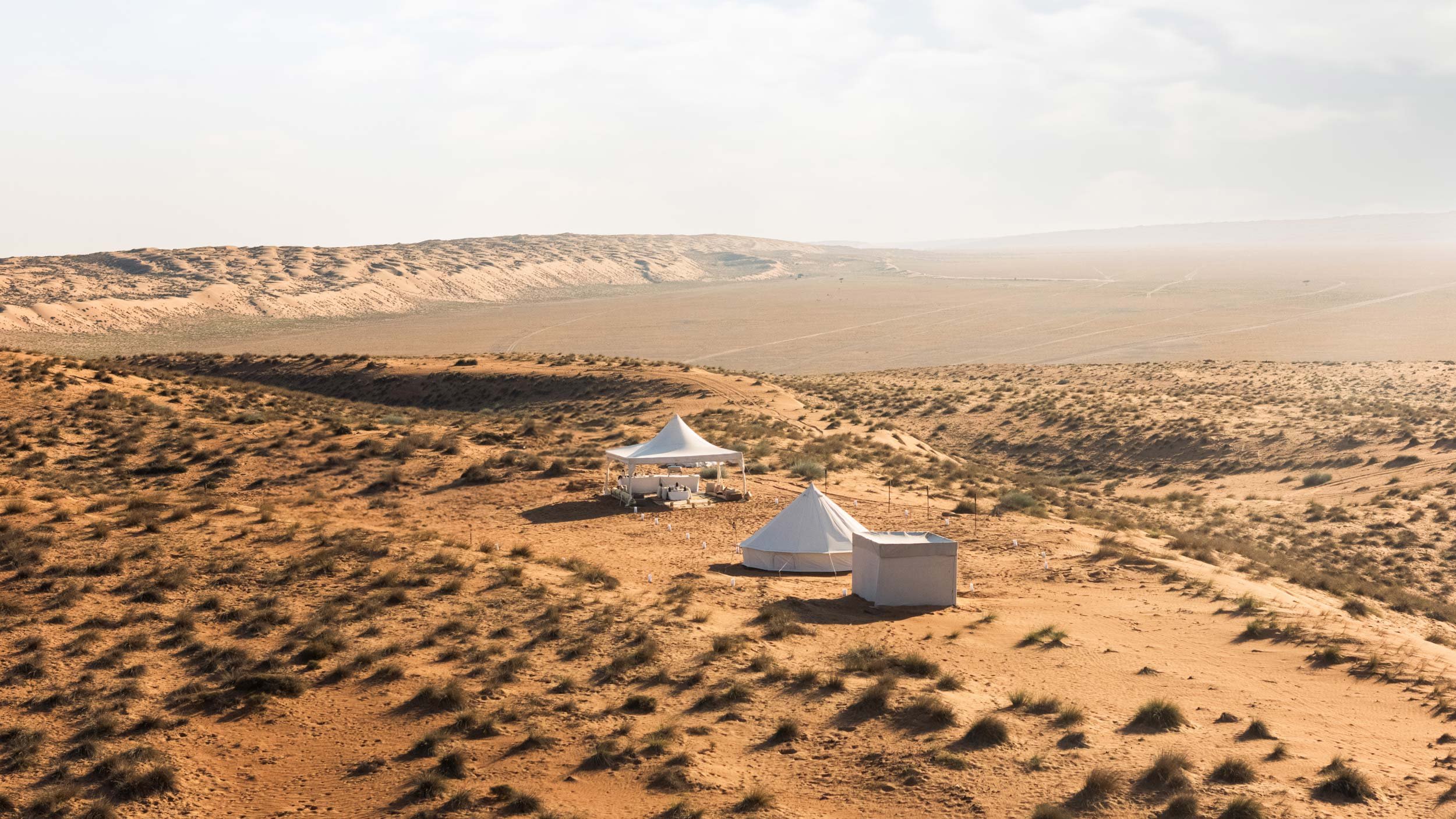 A desert landscape with sandy dunes and sparse vegetation, featuring a white tent and a small structure, under a cloudy sky in Wahiba Sands - Bidiyah - Oman.