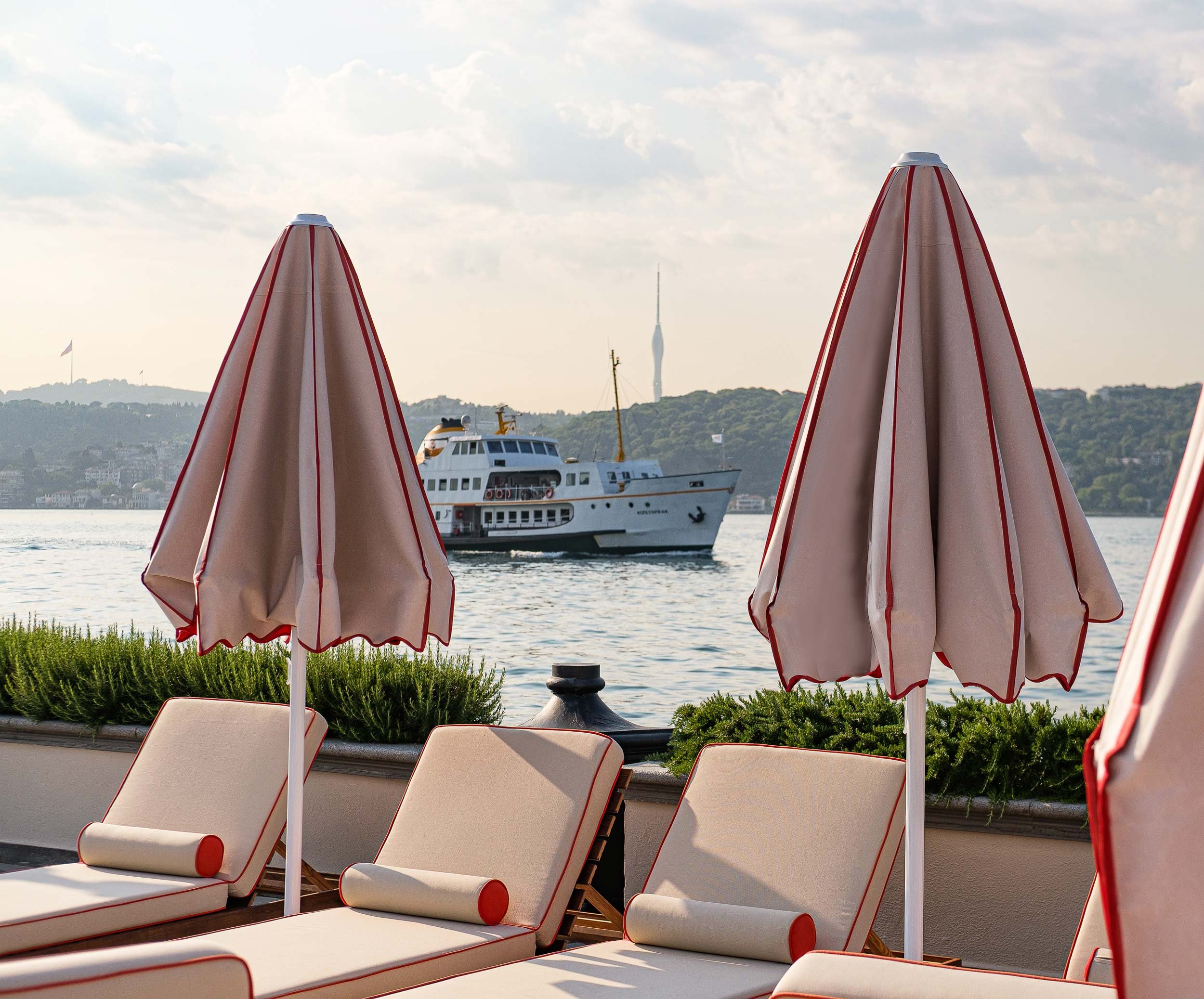 Beach chairs and umbrellas overlooking a river with a boat and distant cityscape, including a tall tower and hill with a flag.