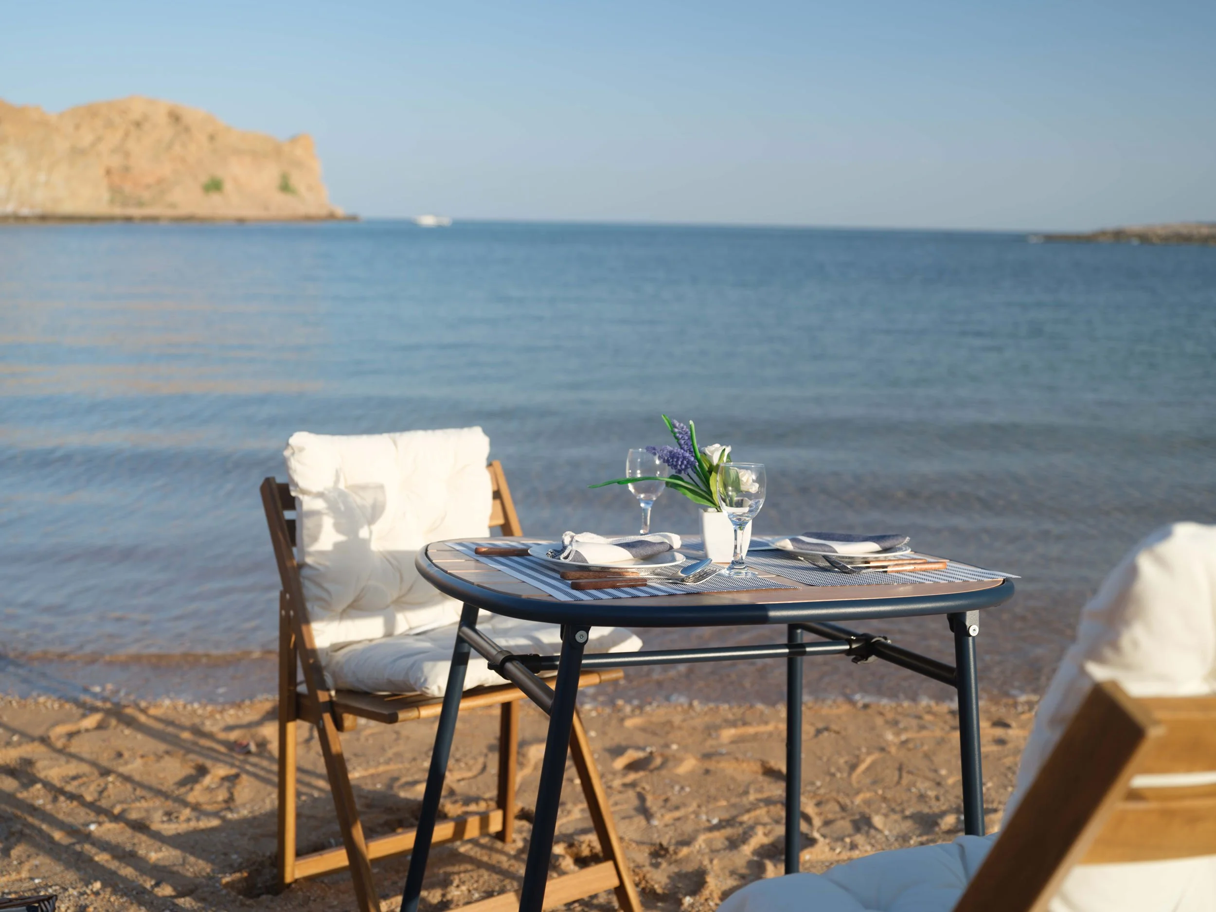 Beachside dining setup with a table, chairs, and a flower centerpiece, overlooking calm ocean waters and rocky cliffs in the background in Bandar al Khiran - Oman.