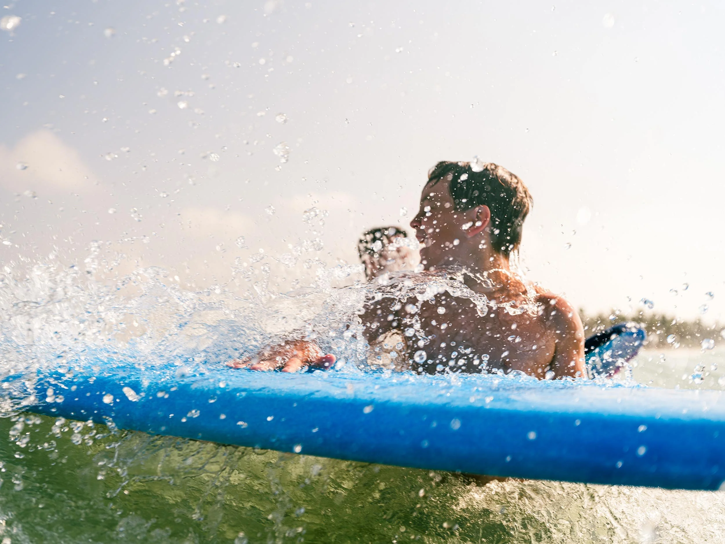 Two young men surfing on a blue paddleboard on the water with splashes of water in the air, with the sun setting in the background.