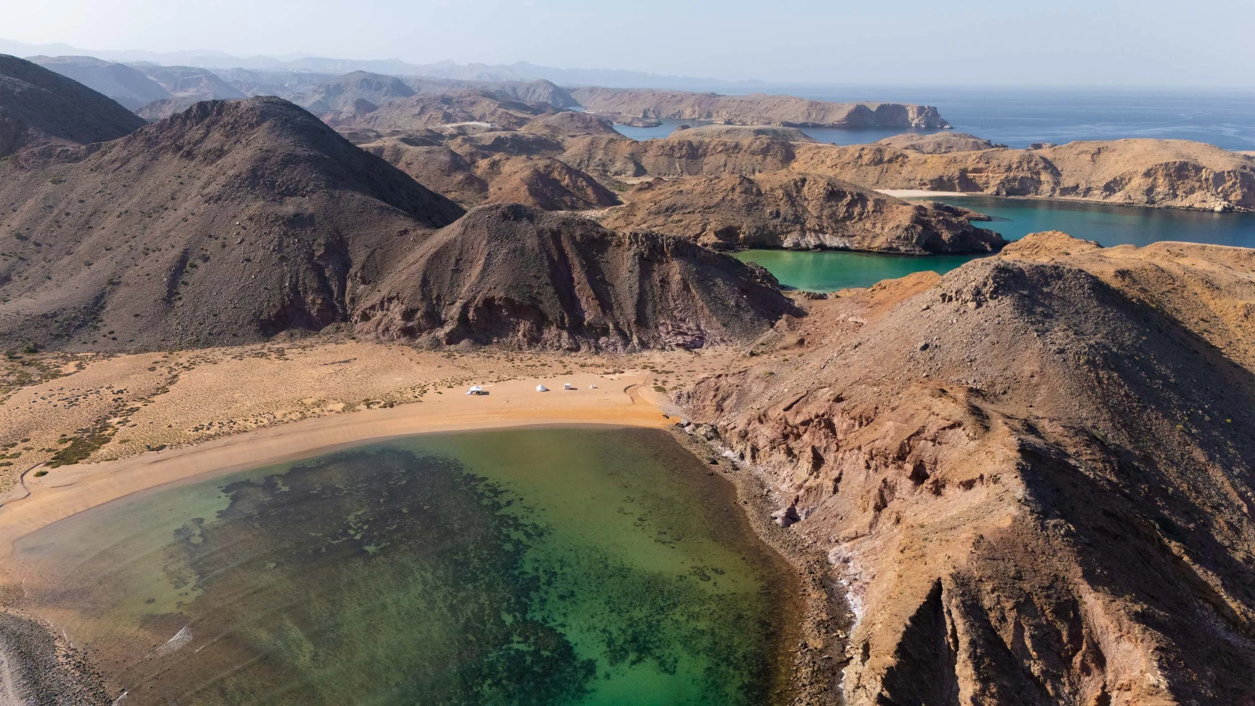 Aerial view of a desert landscape with multiple lakes and mountains, including a small sandy beach area, brown rugged mountains, green and blue lakes, and distant ocean in bandar al khiran - Muscat - Oman.