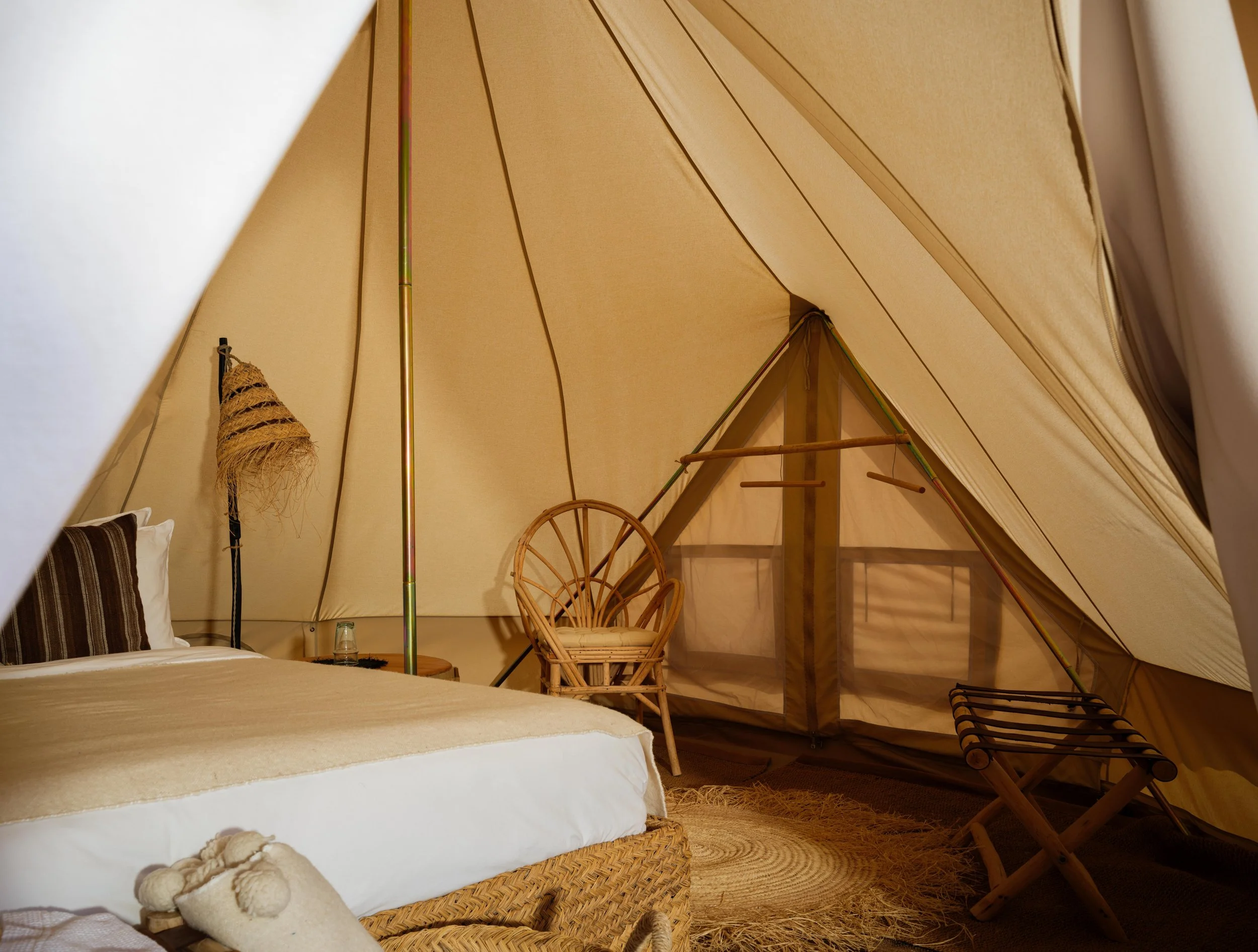 Interior of a glamping tent with a bed, woven chair, wooden stool, lamp, and clothes hangers on a stand, under beige fabric.