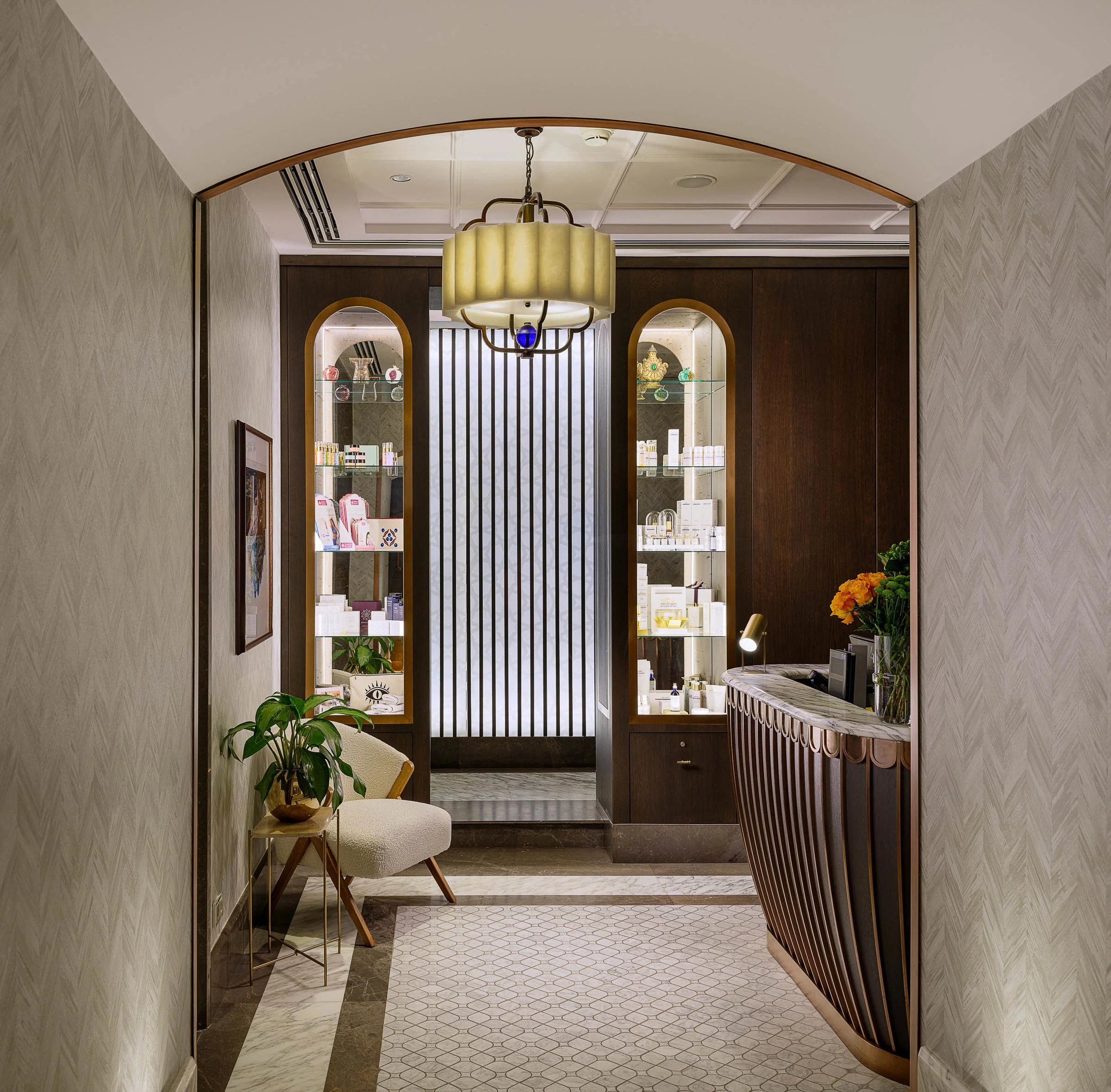 Elegant hotel reception area with a curved wooden front desk, a modern pendant light, decorative shelves with products, a beige chair with a plant, and flowers on the desk, featuring a patterned rug and textured wallpaper.
