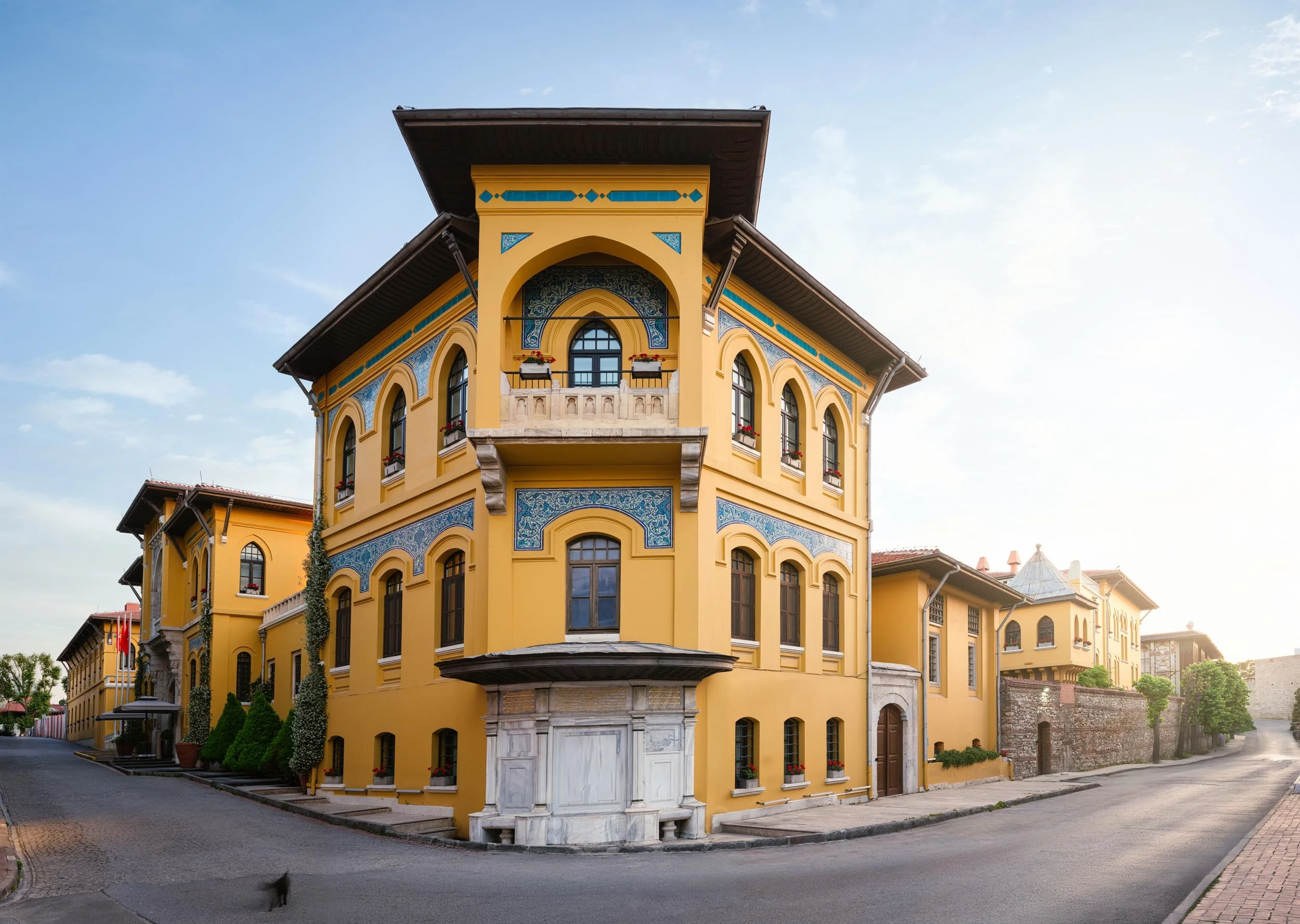 Four Seasons Istanbul Hotel at Sultanahmet which situated in a heritage yellow multi-story building with arched windows and decorative blue accents, situated on a curved street with cobblestone and brick paving, under a partly cloudy sky.
