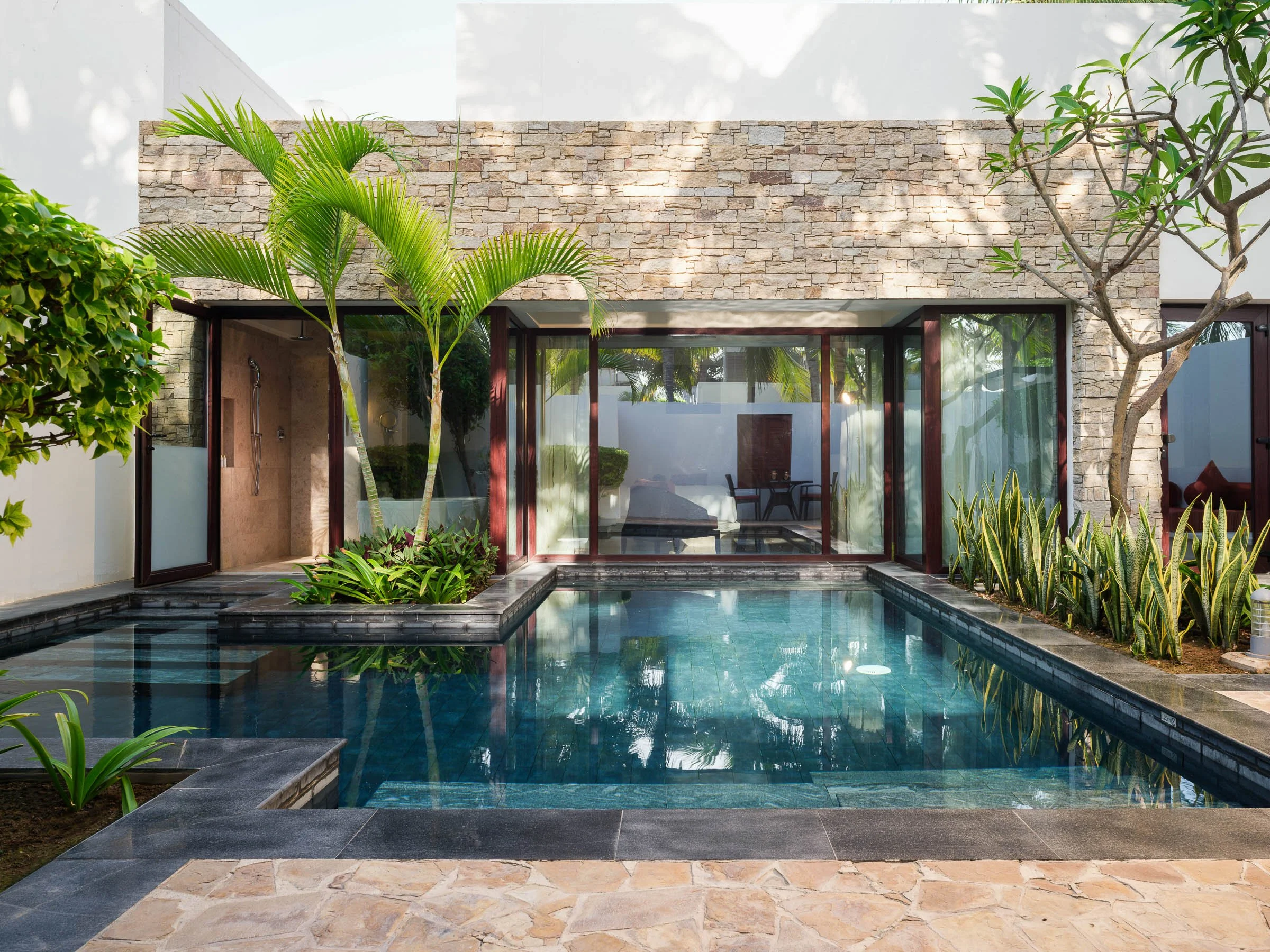 A modern backyard pool area with clear water, surrounded by plants, trees, and a stone patio, with a house featuring a stone wall and glass sliding doors in the background.