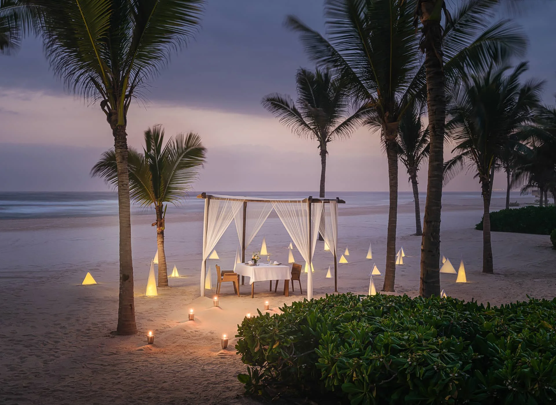 Beach scene at dusk with a decorated dining setup under a canopy, surrounded by illuminated cone-shaped lights and palm trees.