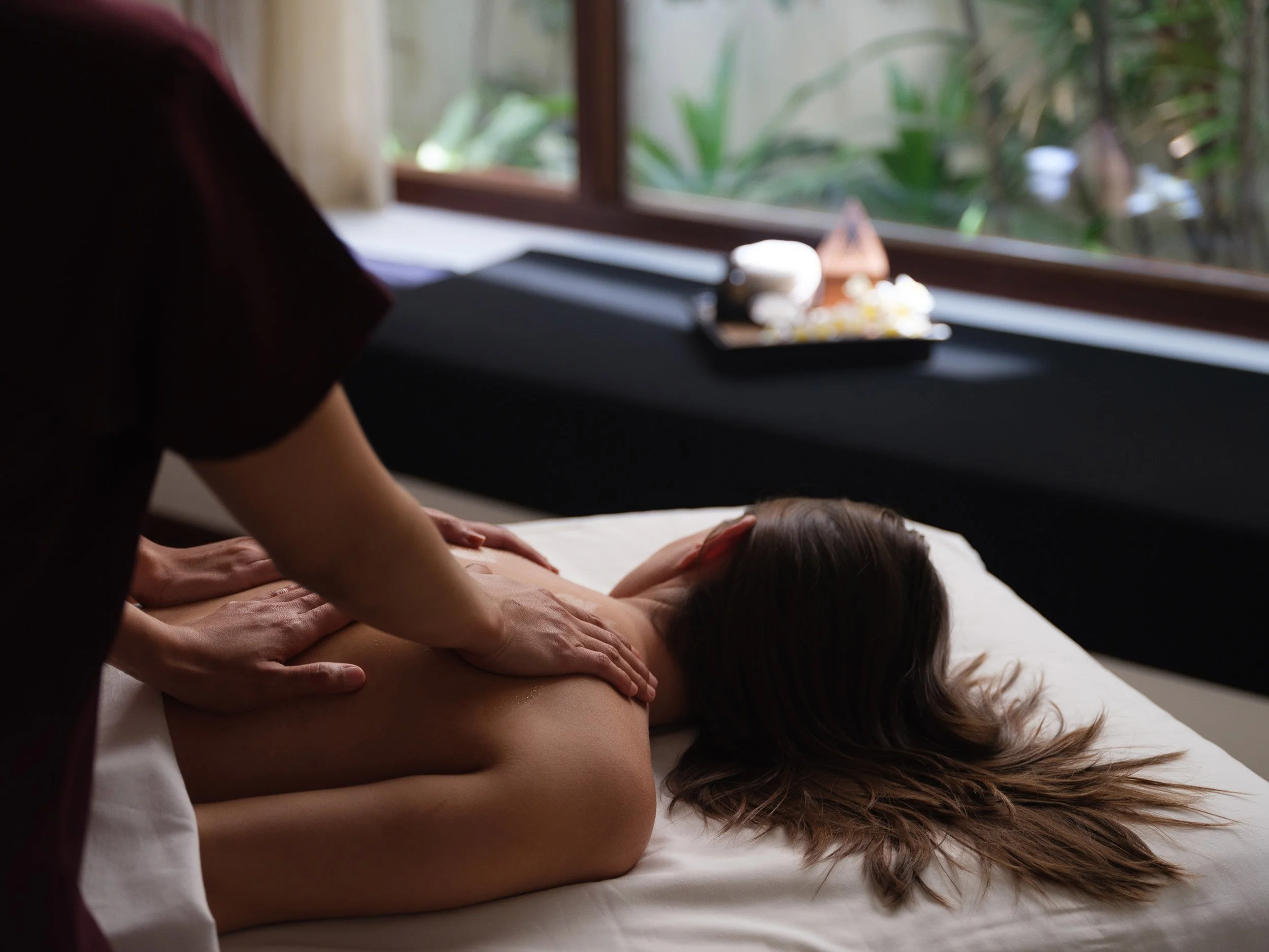 A woman receiving a back massage from a massage therapist in a spa room with a window showing lush green plants outside.