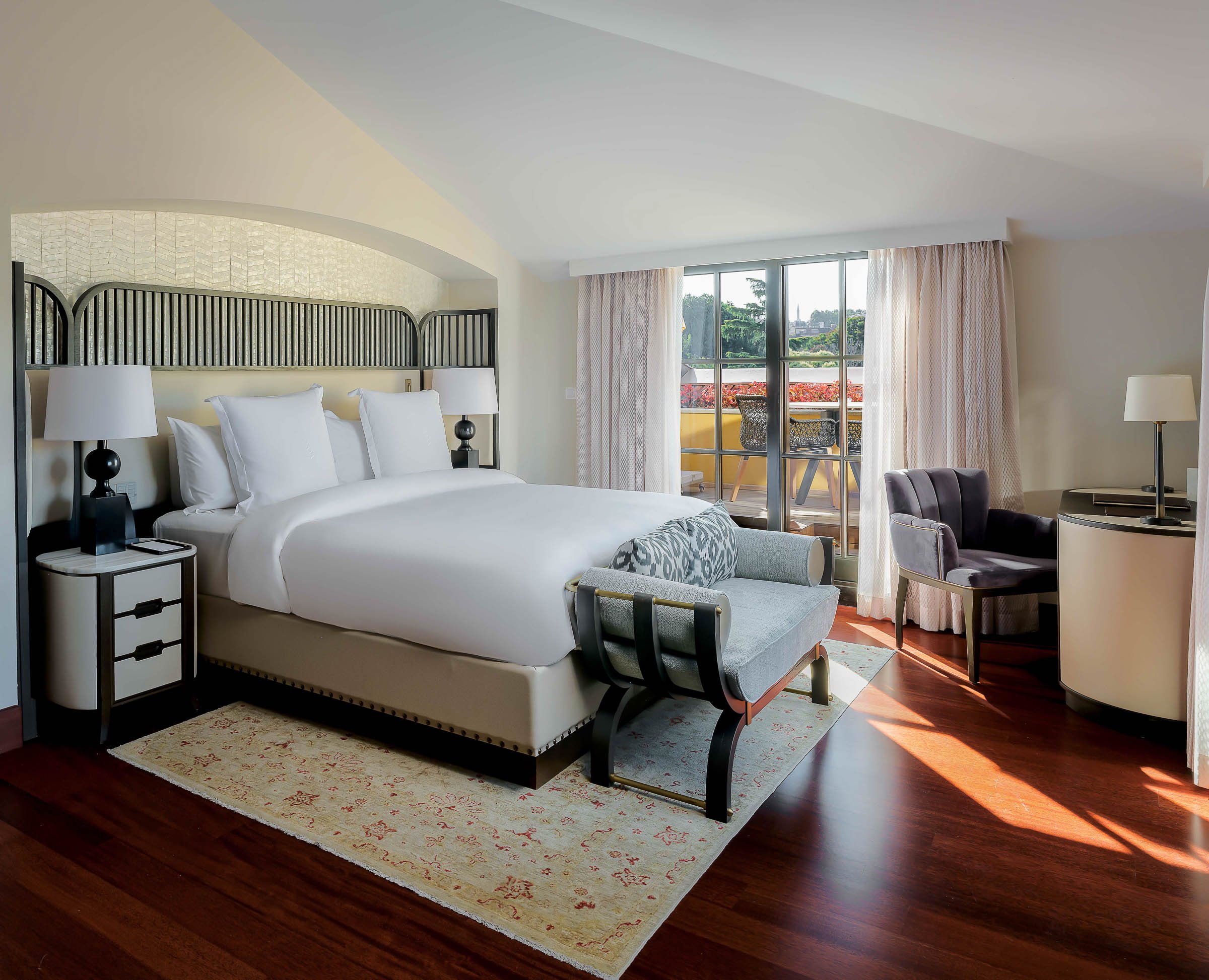 A modern hotel room with a large bed, nightstands with lamps, a balcony with outdoor seating, a desk with a chair, and large windows with curtains, sunlight streaming in.