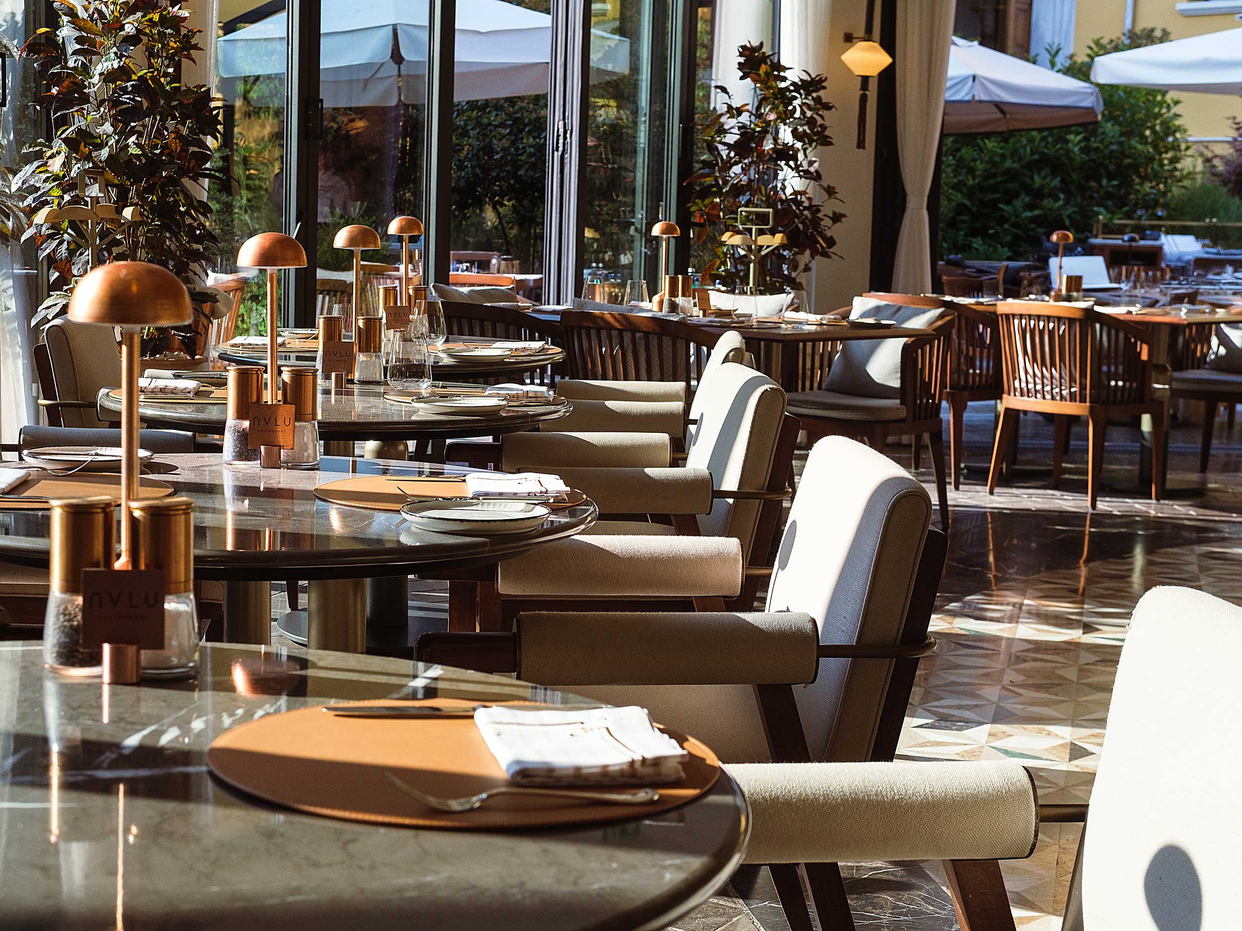 Indoor restaurant with tables set with plates, glasses, and cutlery, illuminated by sunlight through large glass windows, surrounded by plants and outdoor seating visible outside.