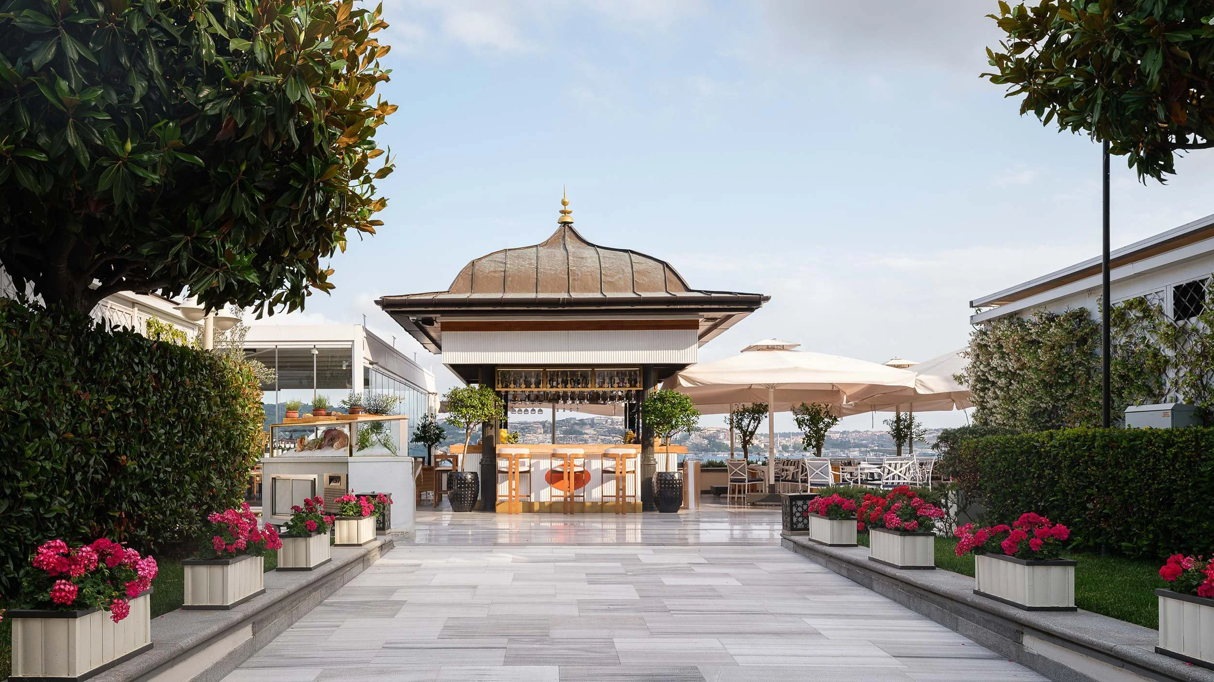An outdoor rooftop bar with a central hut-like structure, surrounded by potted pink flowers and green bushes, with tables and umbrellas in the background under a clear sky.