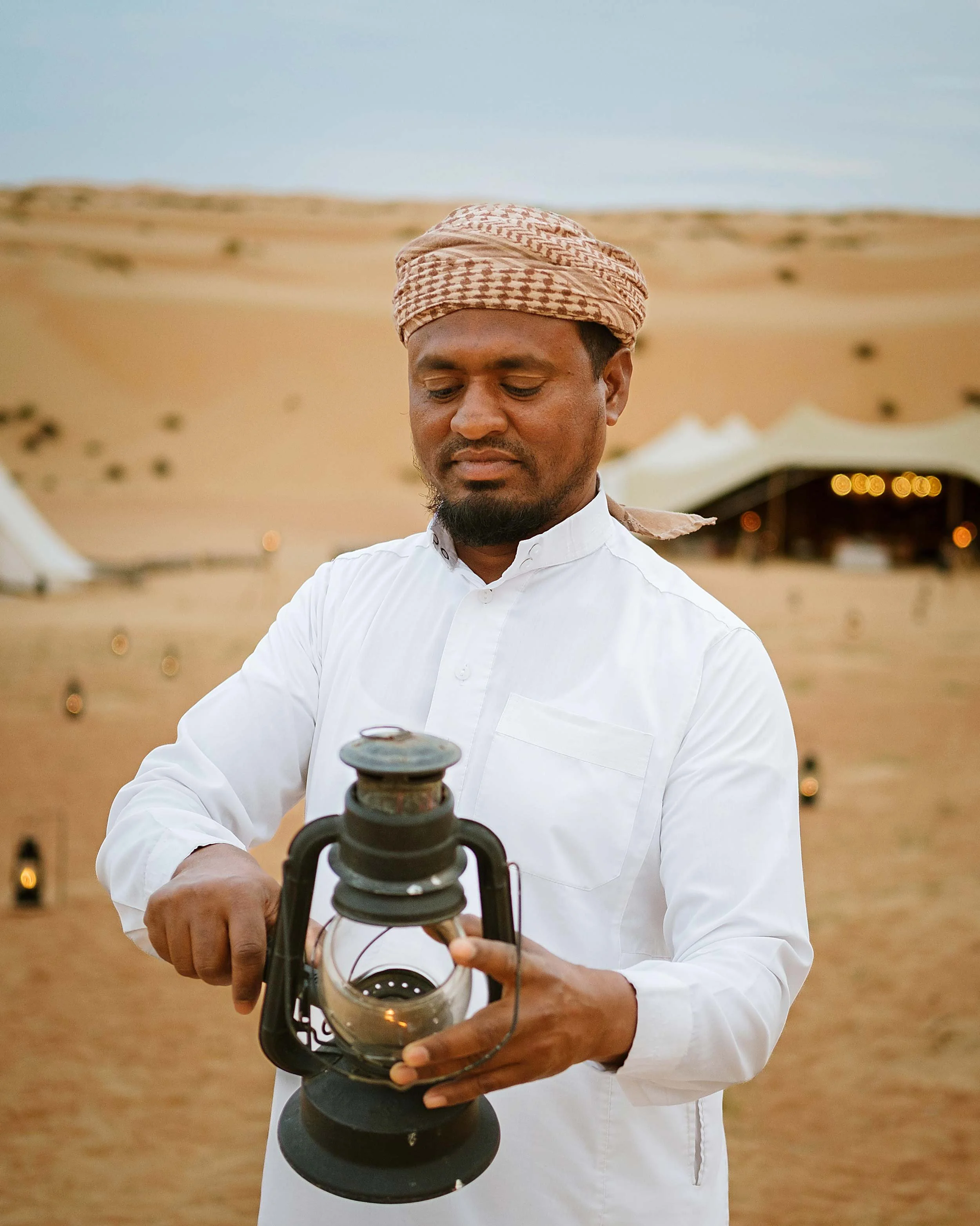 A man wearing a white shirt and a checked headscarf holds an old-fashioned oil lantern in a desert landscape.