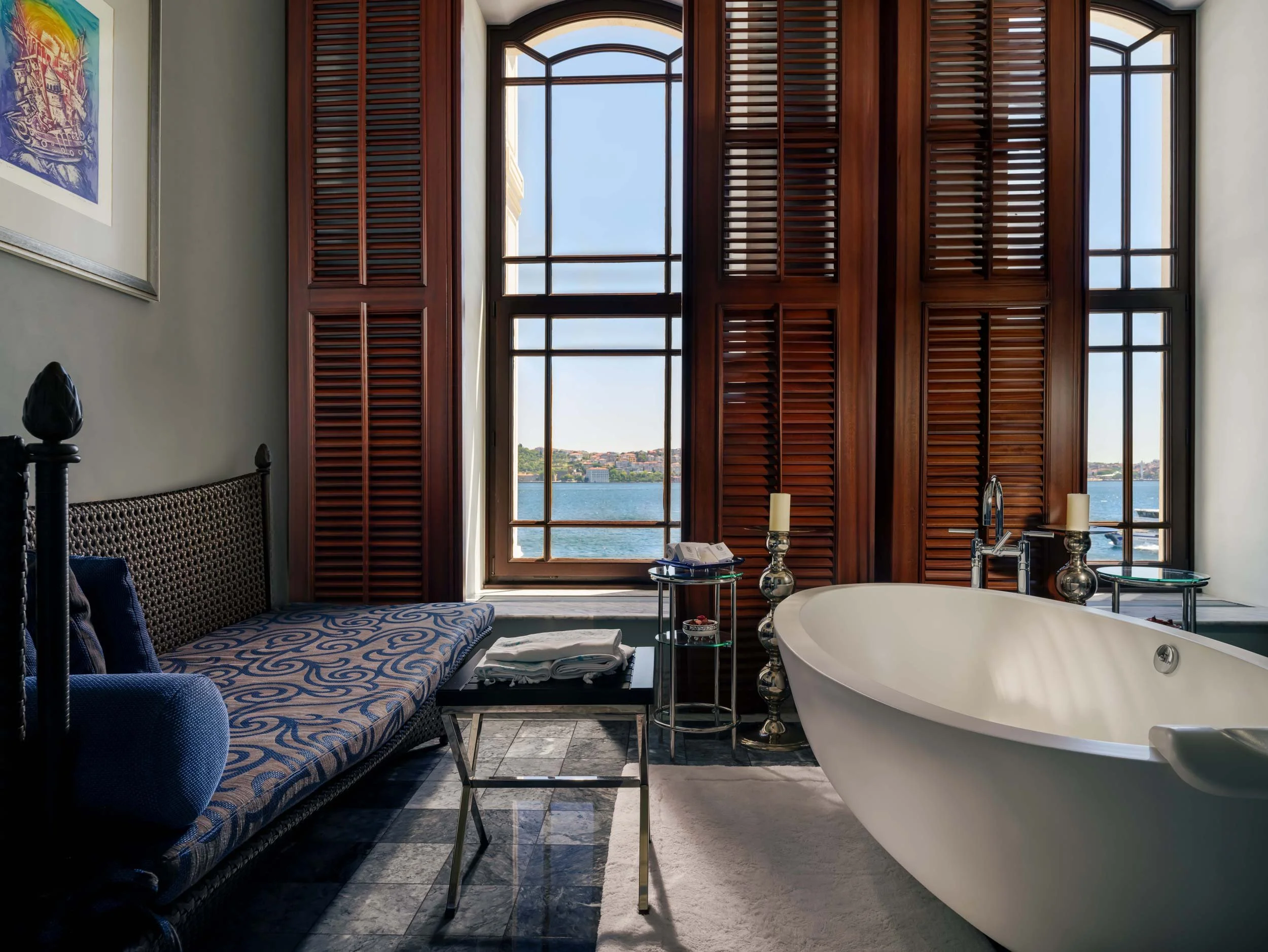 Luxury bathroom with a freestanding bathtub, large windows with wooden shutters overlooking a body of water, a patterned bench with pillows, small tables with candles, and folded towels at Four Seasons Bosphorus Hotel.