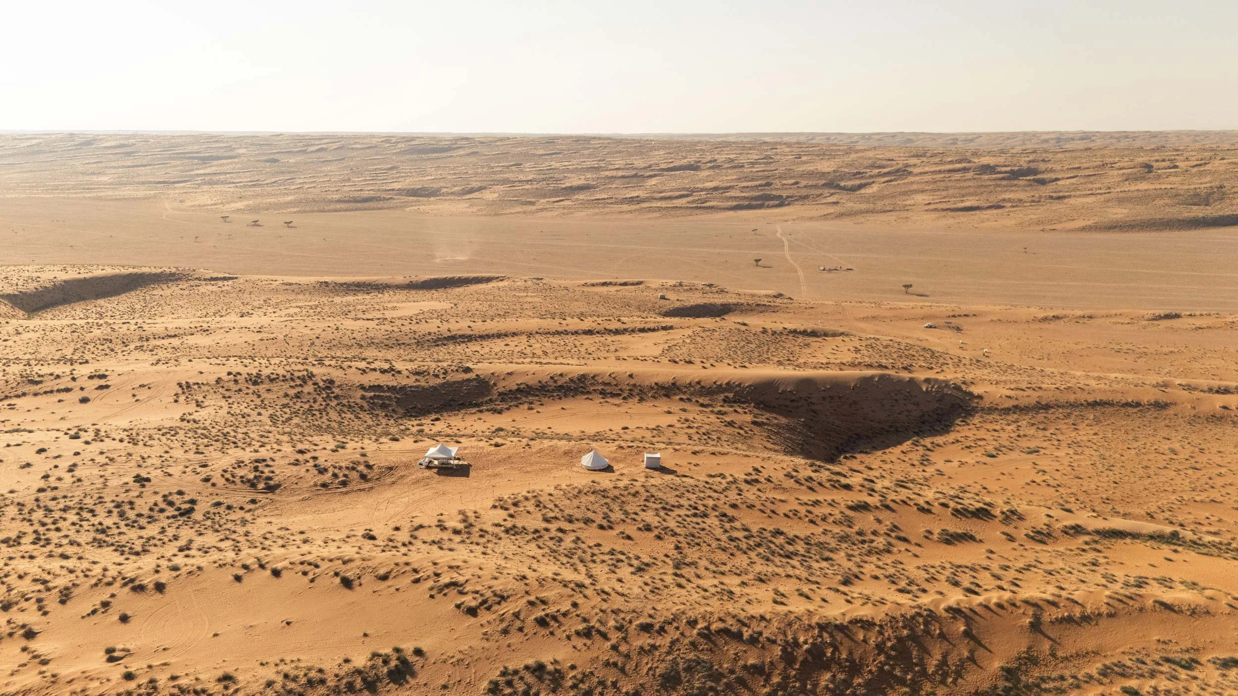 Aerial view of a desert with sparse vegetation, a few tents, and some vehicles, surrounded by sand dunes and flat terrain in empty quarter - Wahiba Sands.