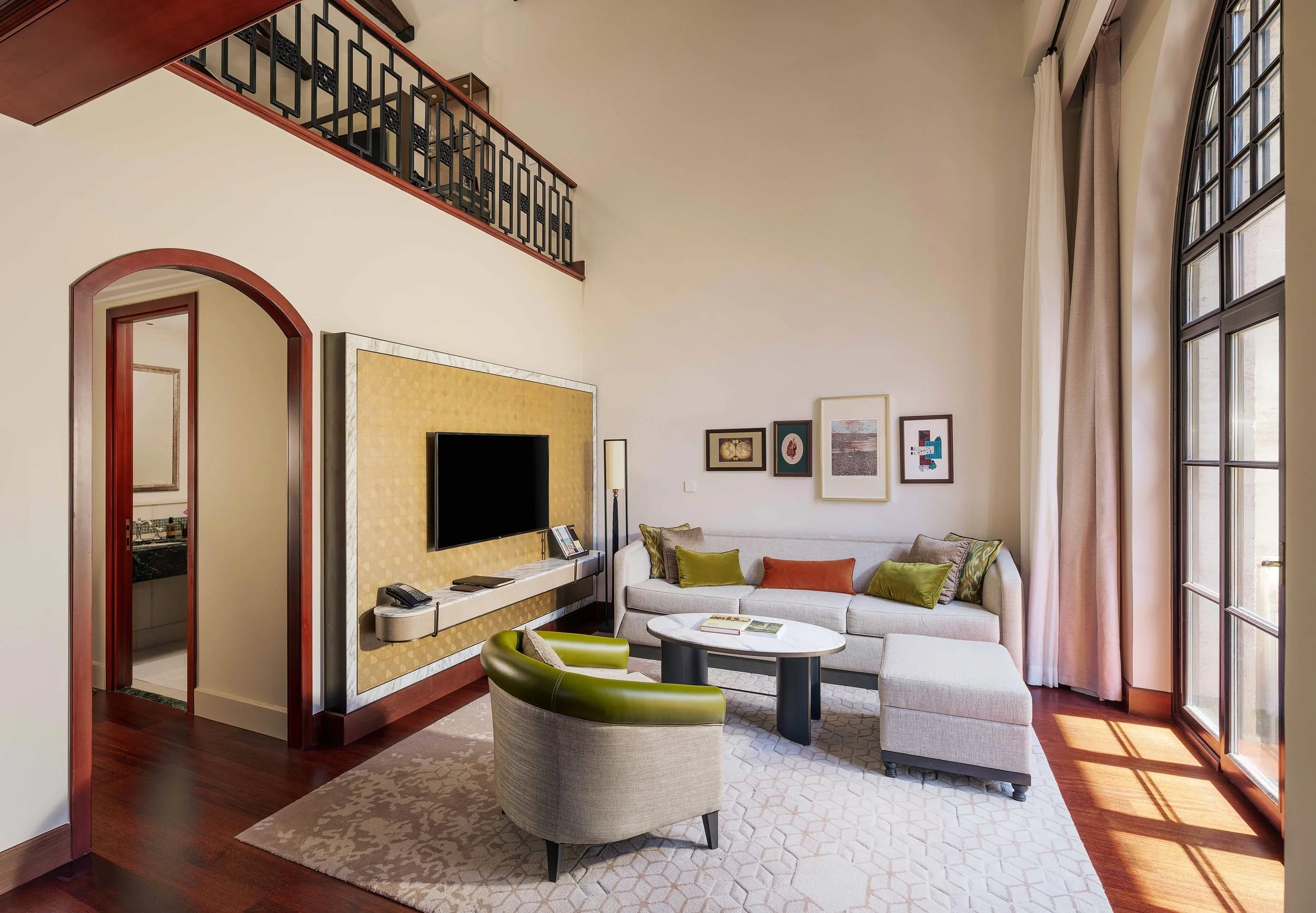 Living room with beige sofa, green and orange pillows, a round marble coffee table, an upholstered green armchair, a large window with curtains, framed artwork on the wall, a wall-mounted TV, and a partially visible bathroom.