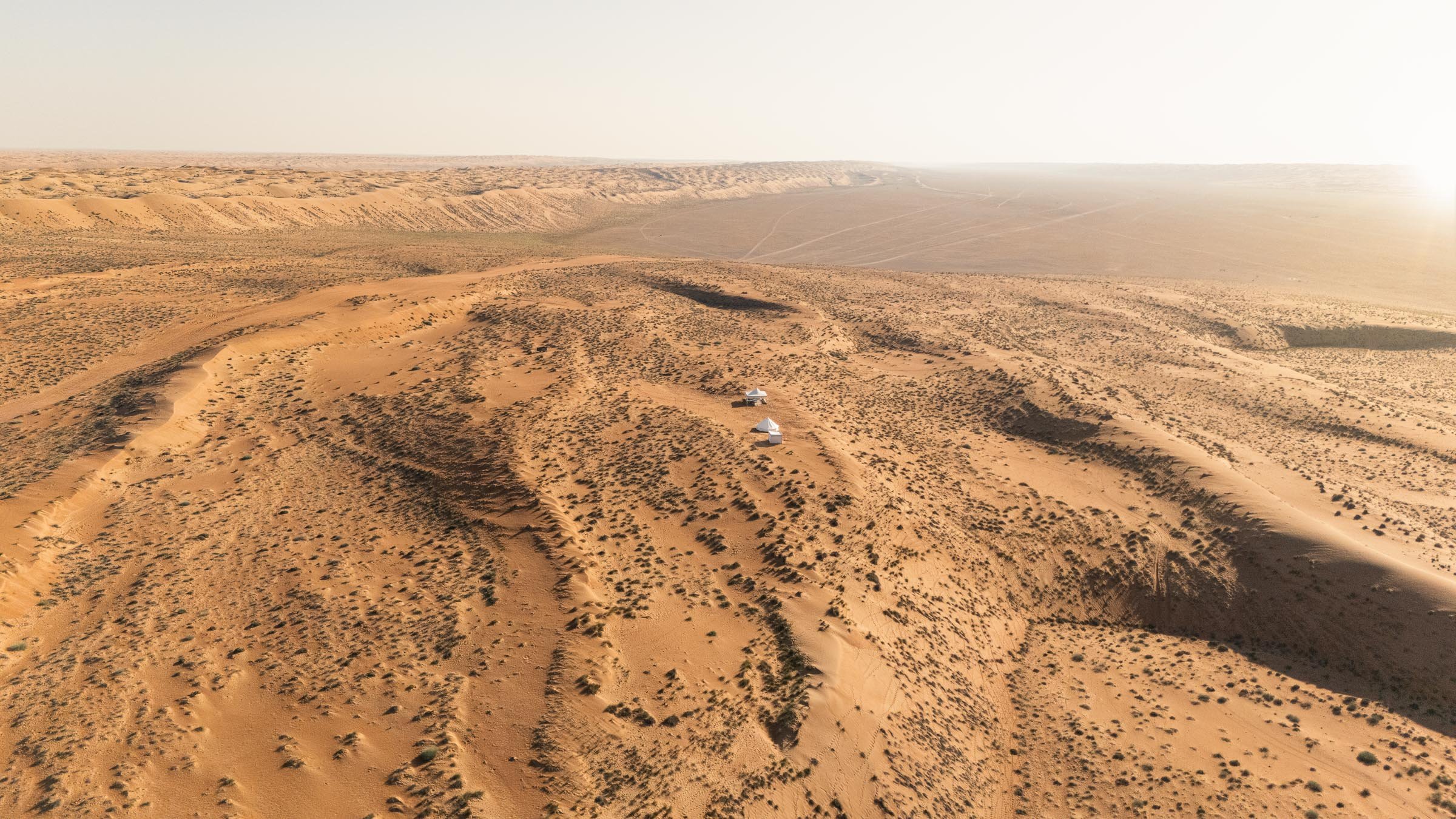 Aerial view of a desert landscape with sand dunes and sparse vegetation, featuring two small white structures in the center.