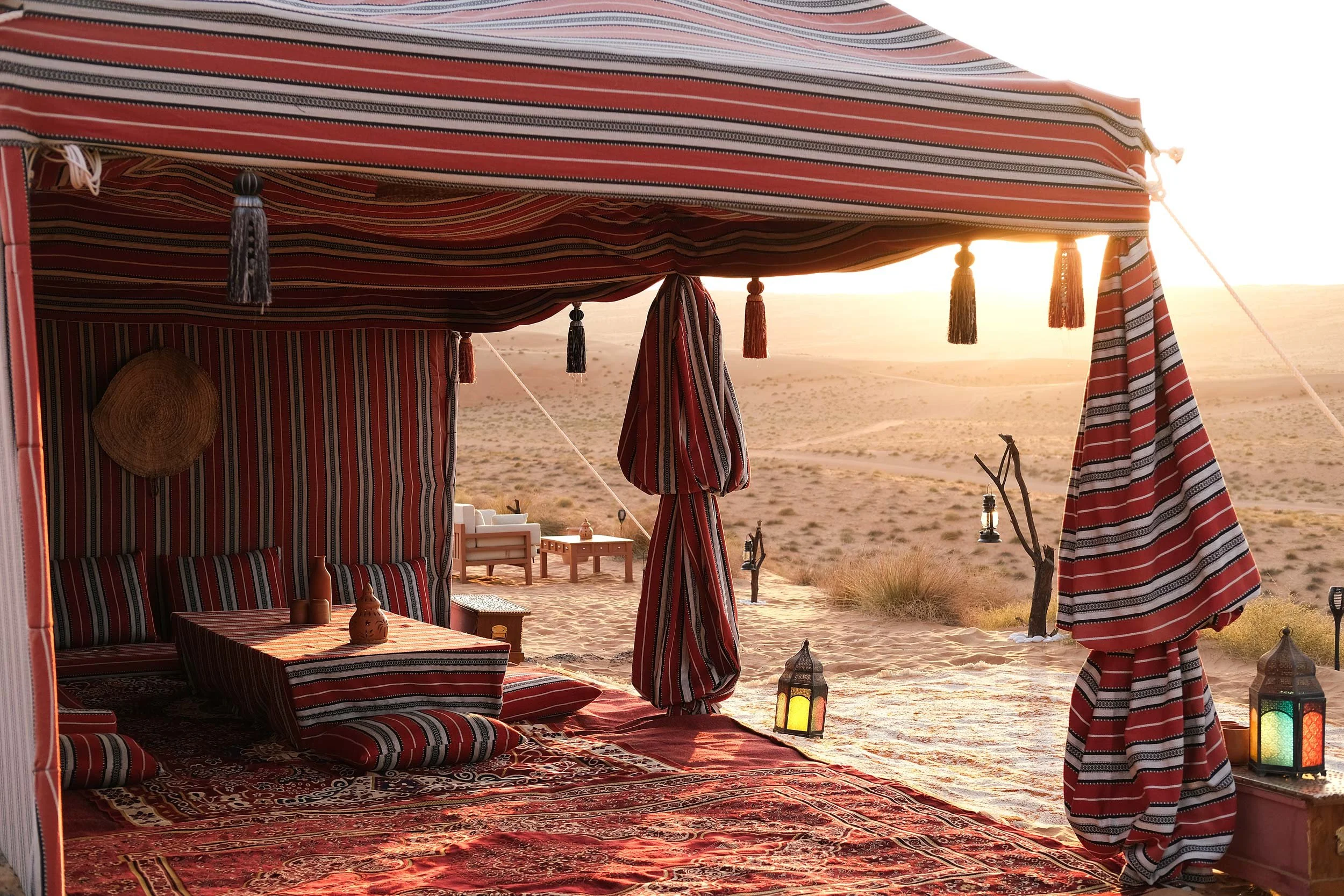 A desert scene viewed from inside a shaded tent with red, black, and white striped fabric, featuring Moroccan-style lanterns, cushions, a low table, and outdoor seating, illuminated by the sunset in empty quarter Oman.