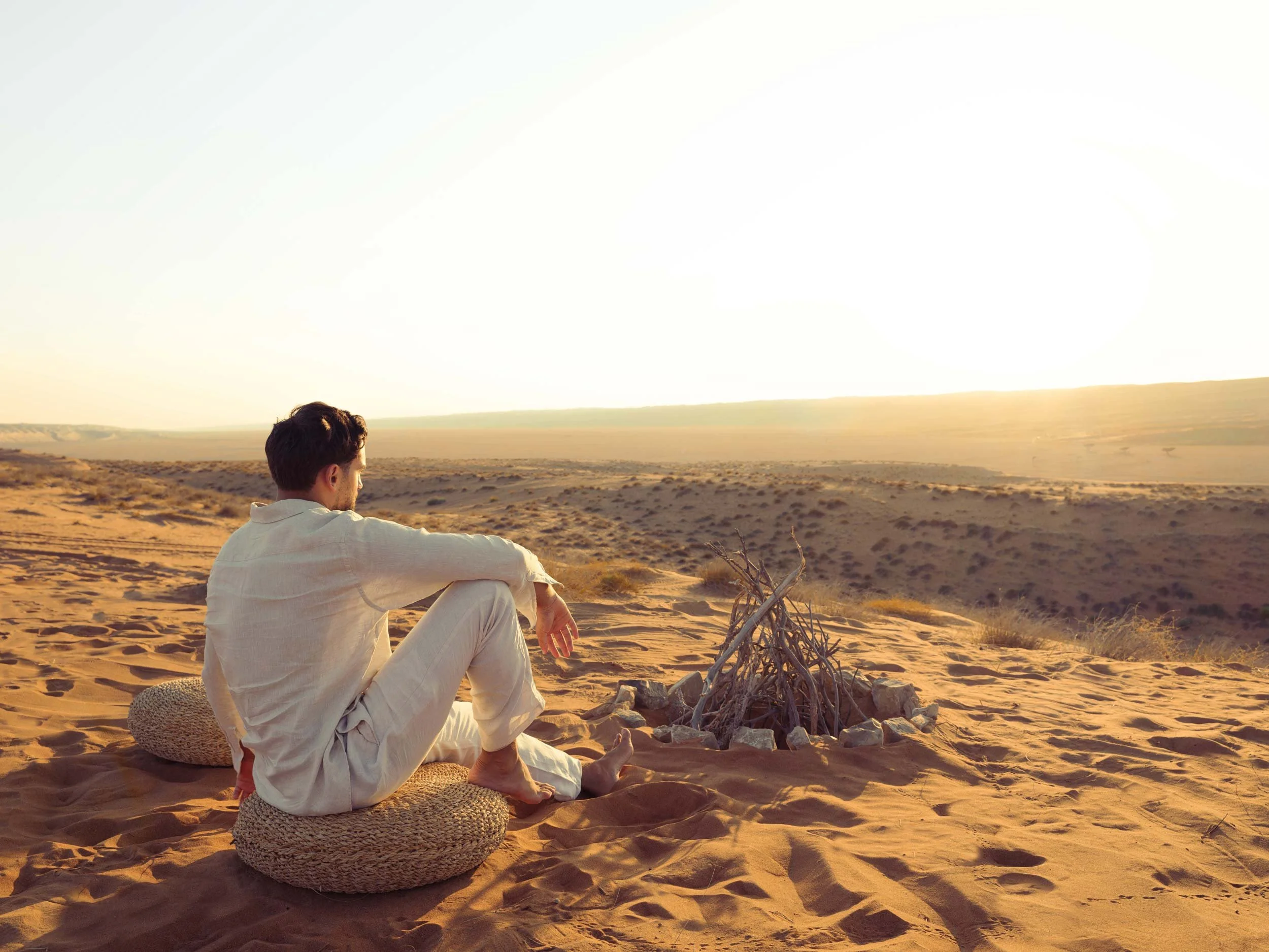A man sitting on a round cushion in a desert, looking at a small campfire made of sticks, with a vast sandy landscape and sunset in the background in Wahiba Sands - Biddiyah.