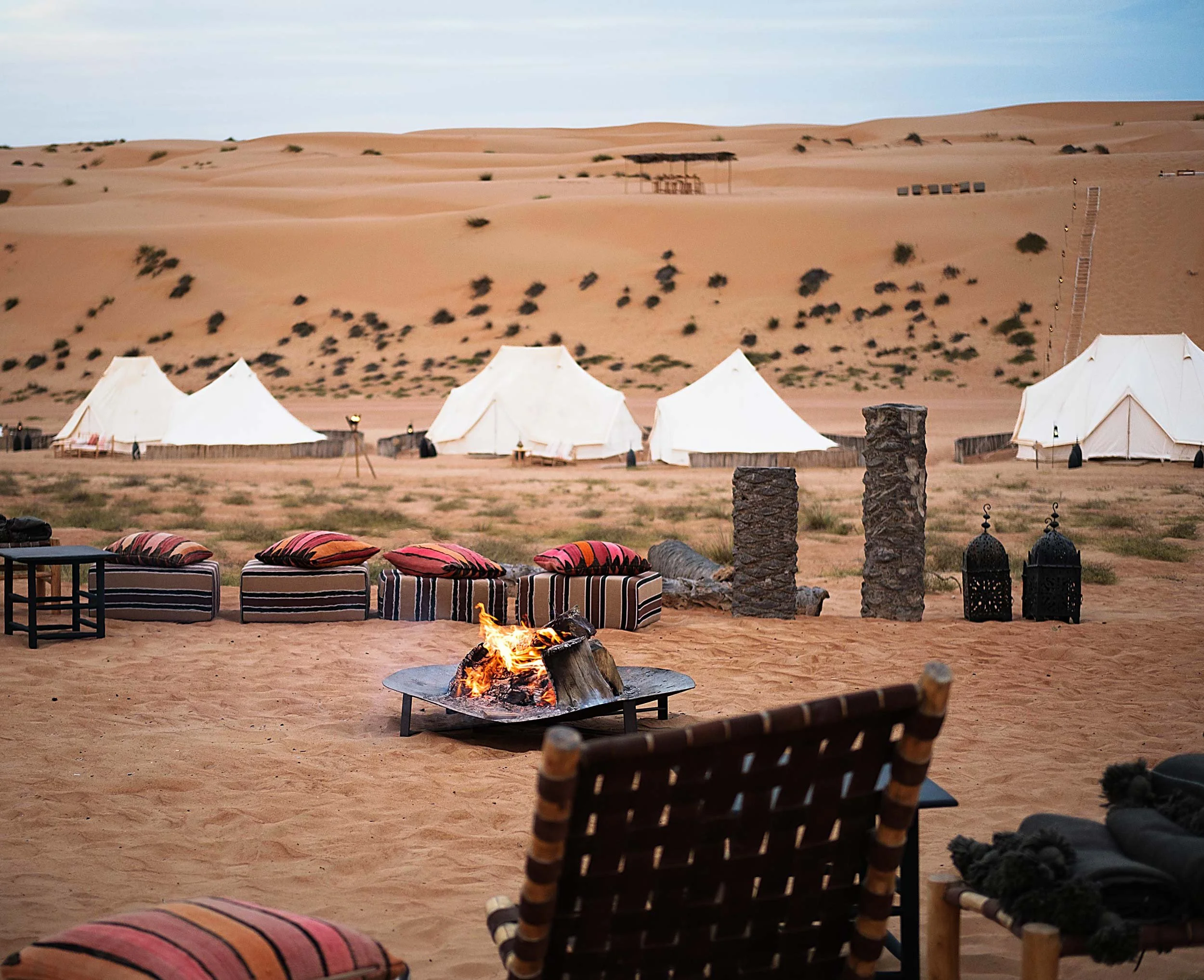 Desert campsite with white tents, colorful cushions on benches, a fire pit, and decorative lanterns set against sand dunes and sparse vegetation.