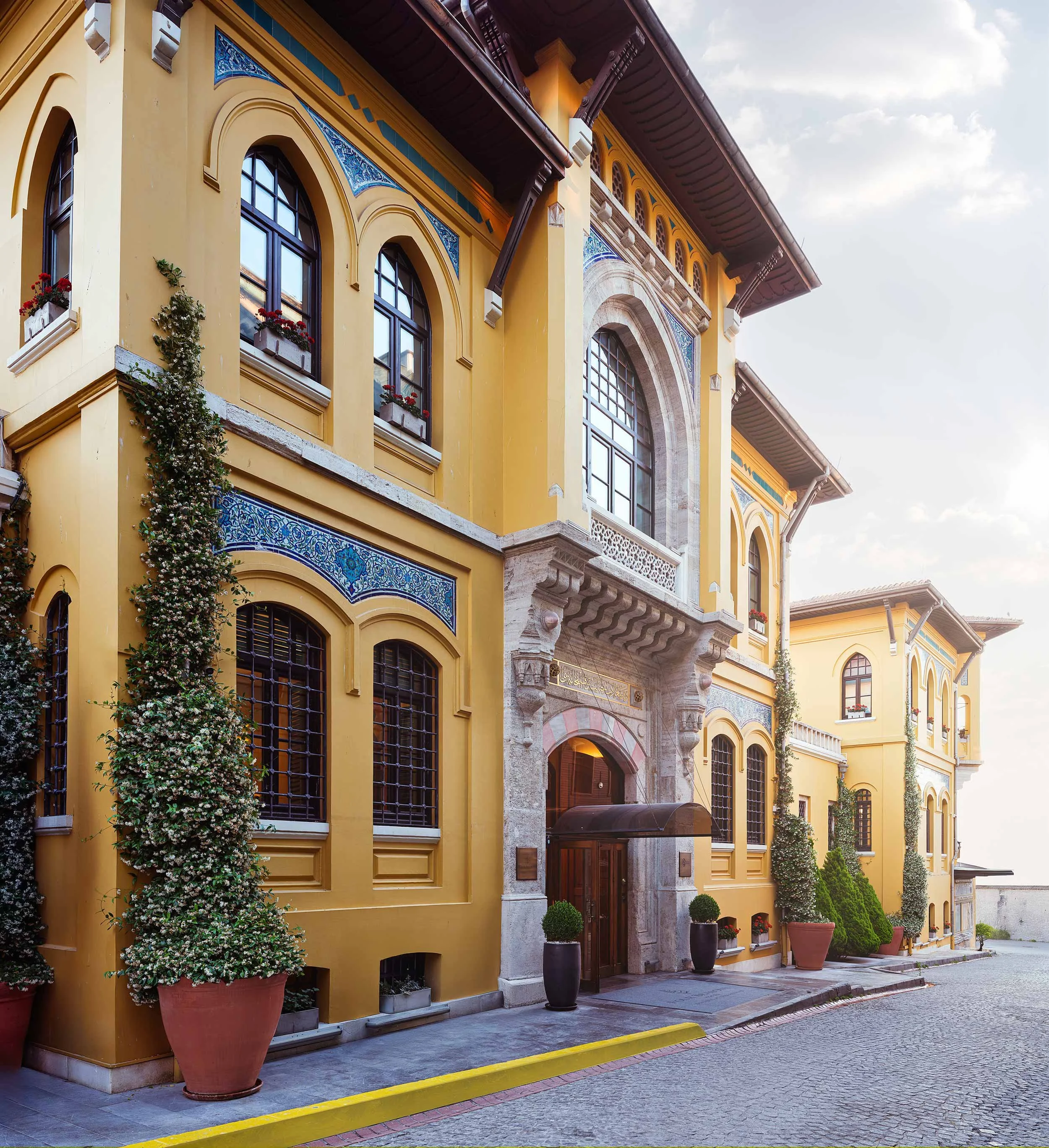 Yellow European-style building with arched windows, blue decorative tiles, potted plants along cobblestone street, and a stone arch entrance, under a partly cloudy sky.