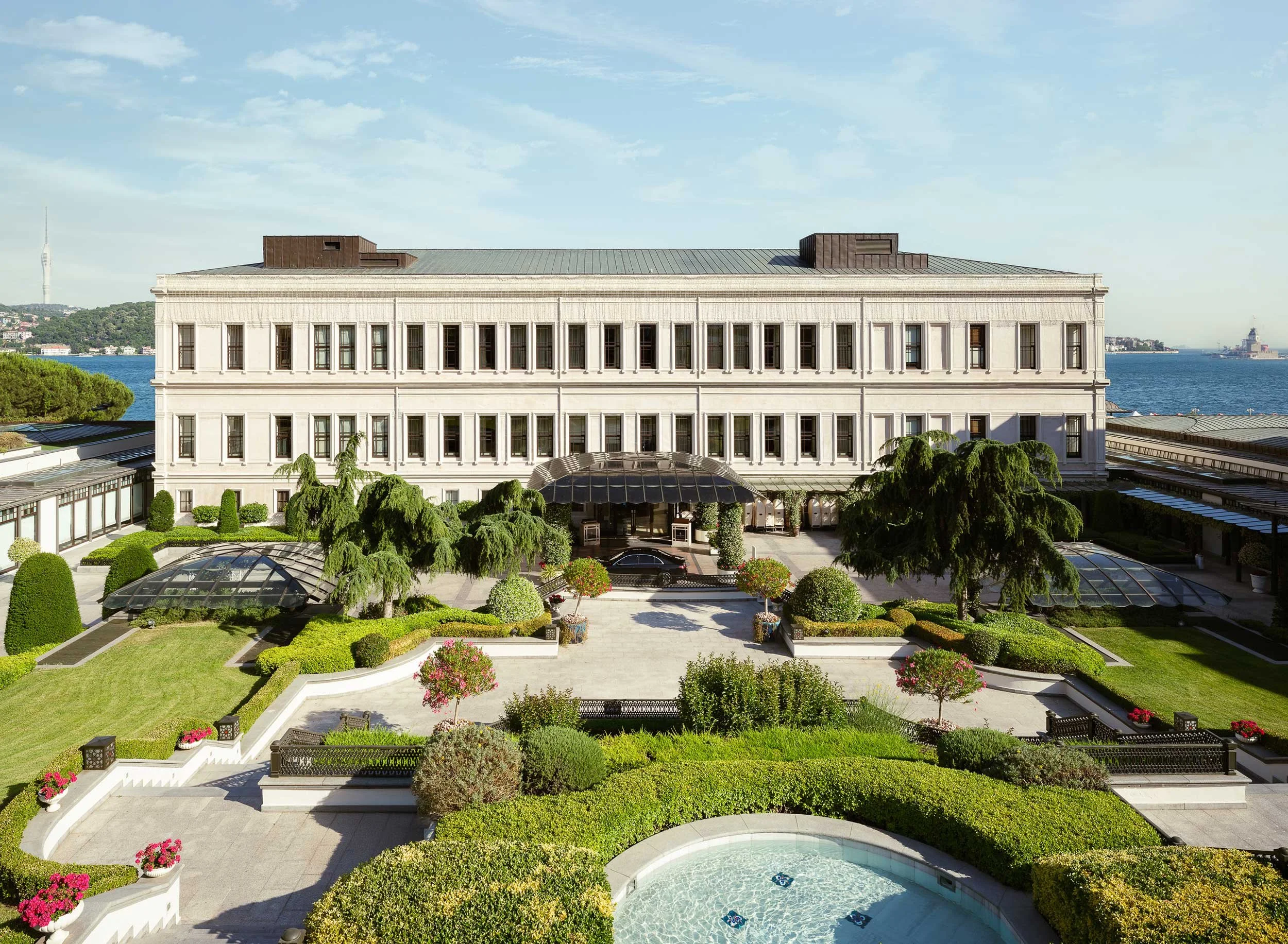 Four Seasons Bosphorus exterior with multiple windows, surrounded by a landscaped garden featuring trees, shrubs, and flower pots, overlooking a body of water with a clear sky overhead.