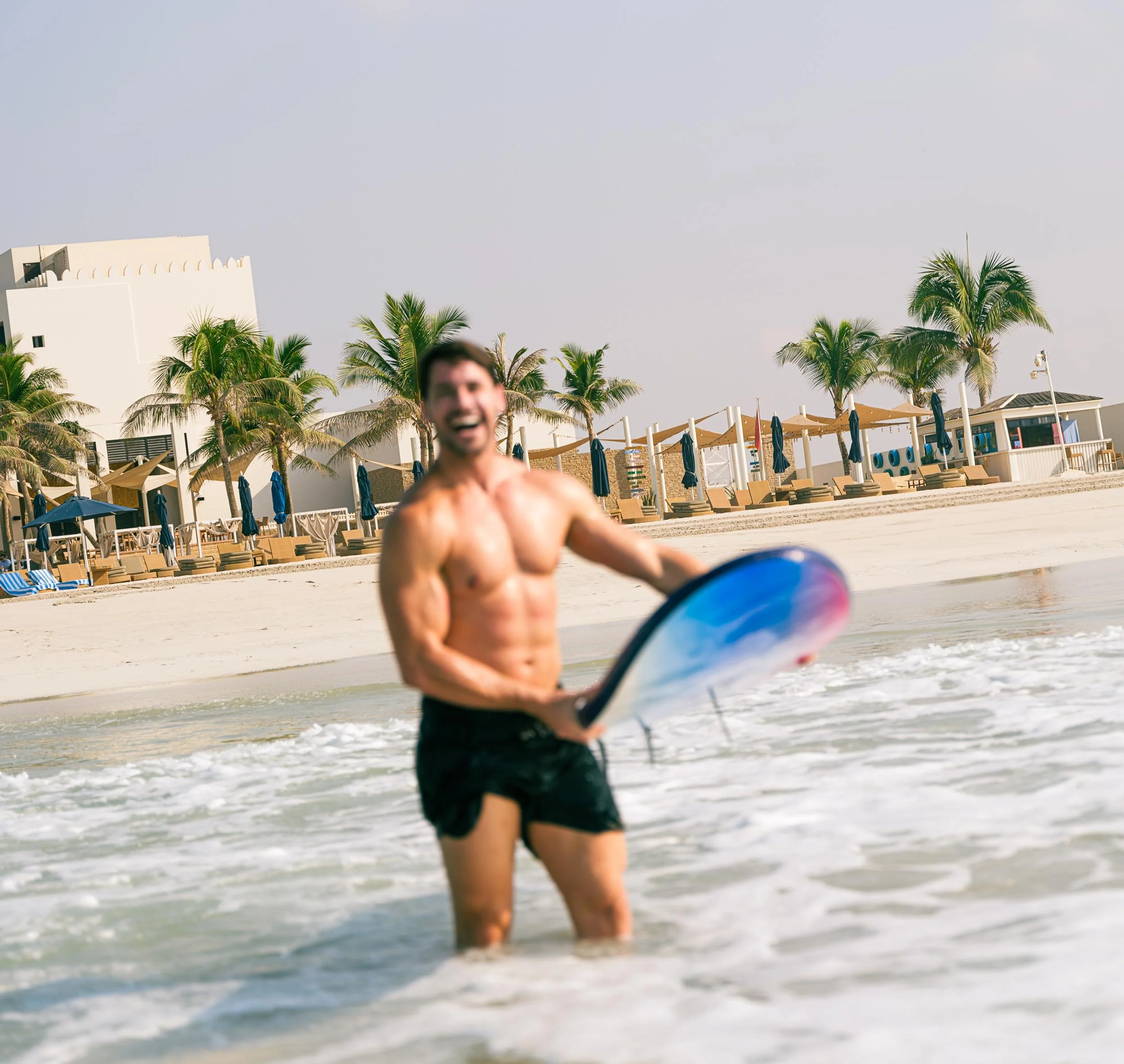A shirtless man with a big smile holding a bodyboard in the shallow surf at a beach with white sand, palm trees, and beach chairs in the background.