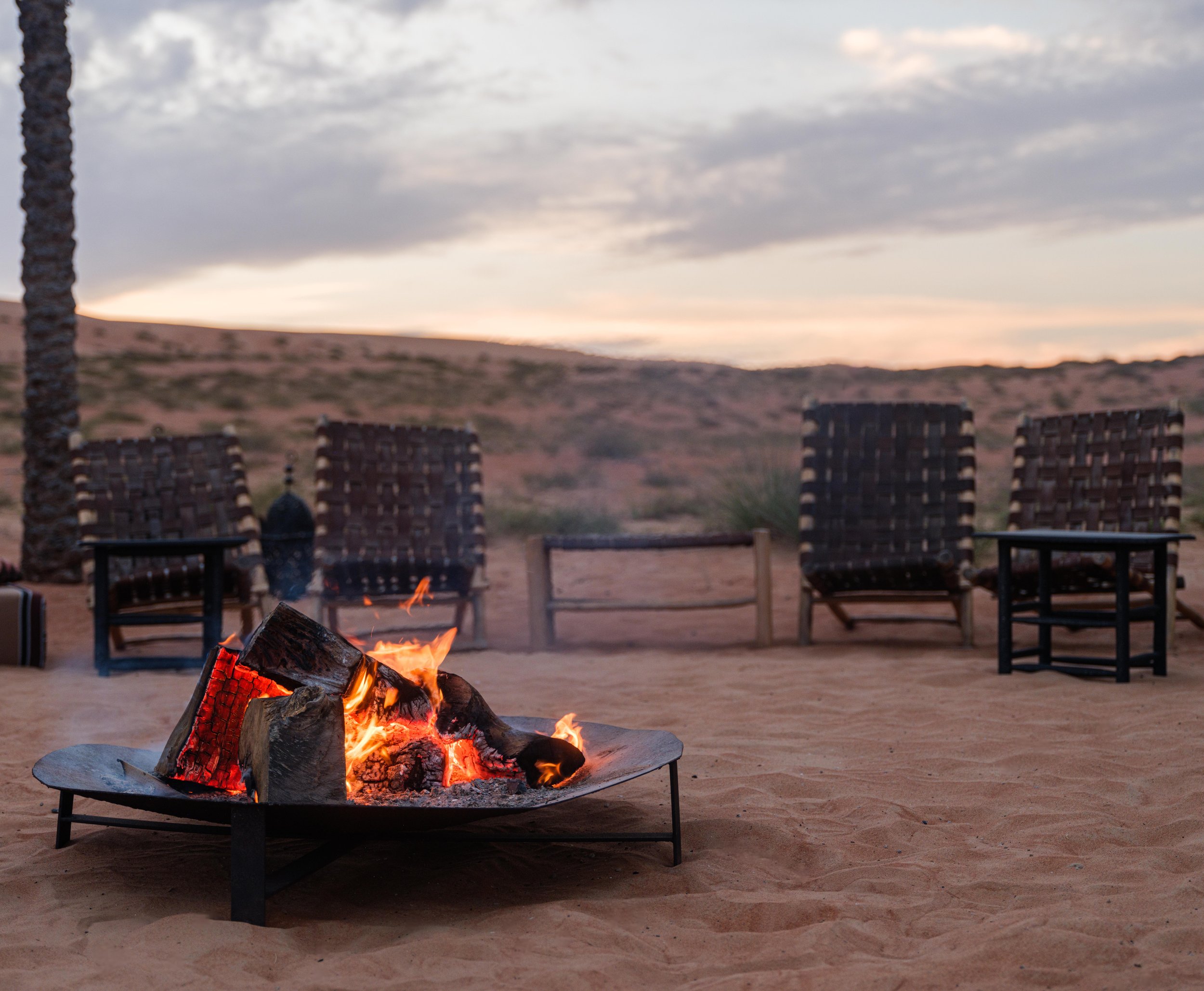 Outdoor fire pit with burning logs, surrounded by chairs on a sandy surface, with a desert landscape background at dusk.