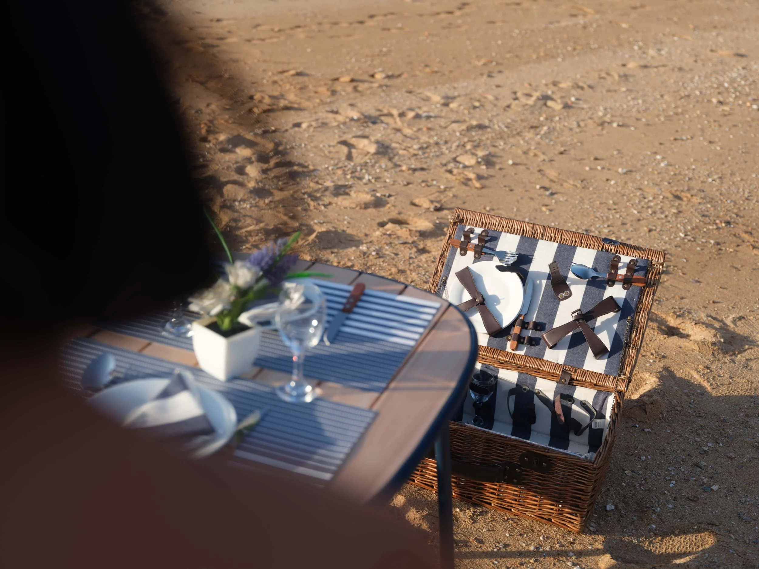 A picnic setup on a sandy beach with a table holding a vase with flowers, plates, glassware, and utensils. A wicker picnic basket with leather straps, filled with various utensils and bottles, is open on the sand nearby.