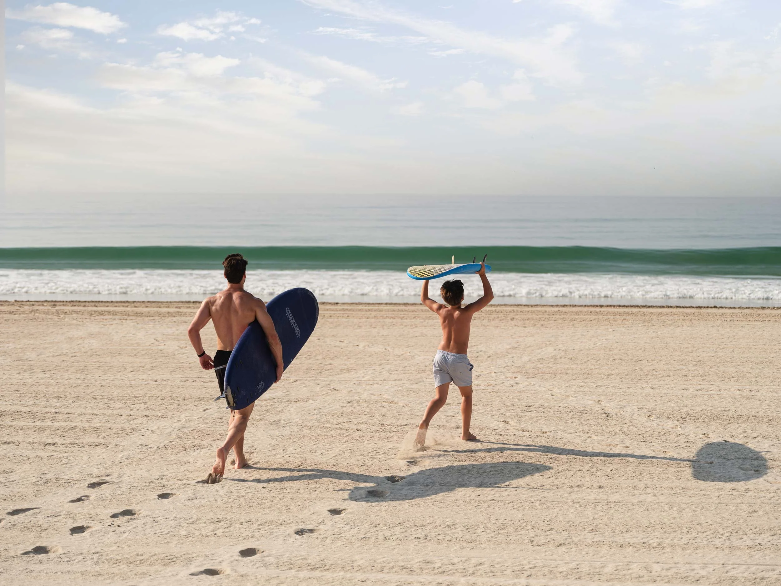 A man and a boy with surfboards walking on the beach towards the ocean, with waves in the background and clouds in the sky.