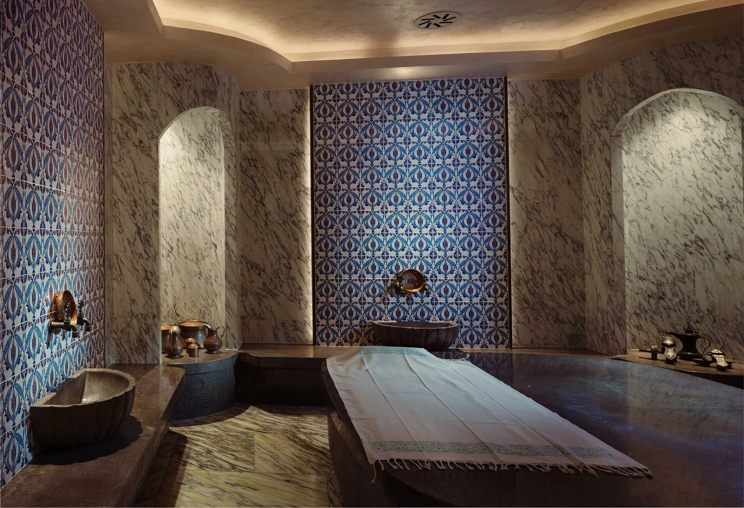 An ornate spa room with dark marble benches, blue patterned tiled walls, marble walls, and a table with a white cloth in the center