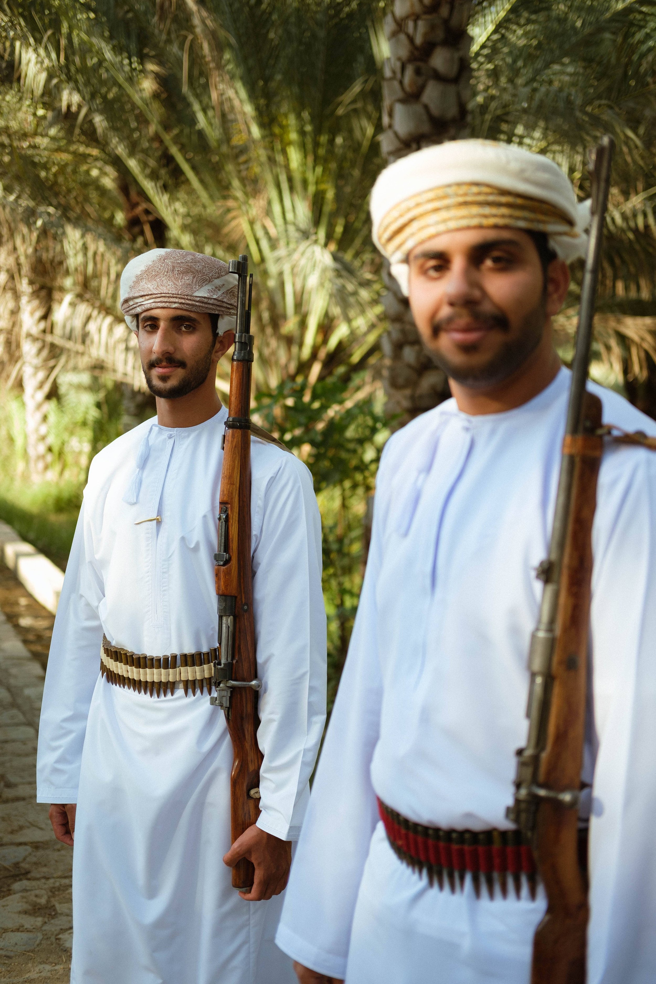 Two men dressed in traditional Middle Eastern clothing, each holding a rifle, standing outdoors among palm trees.