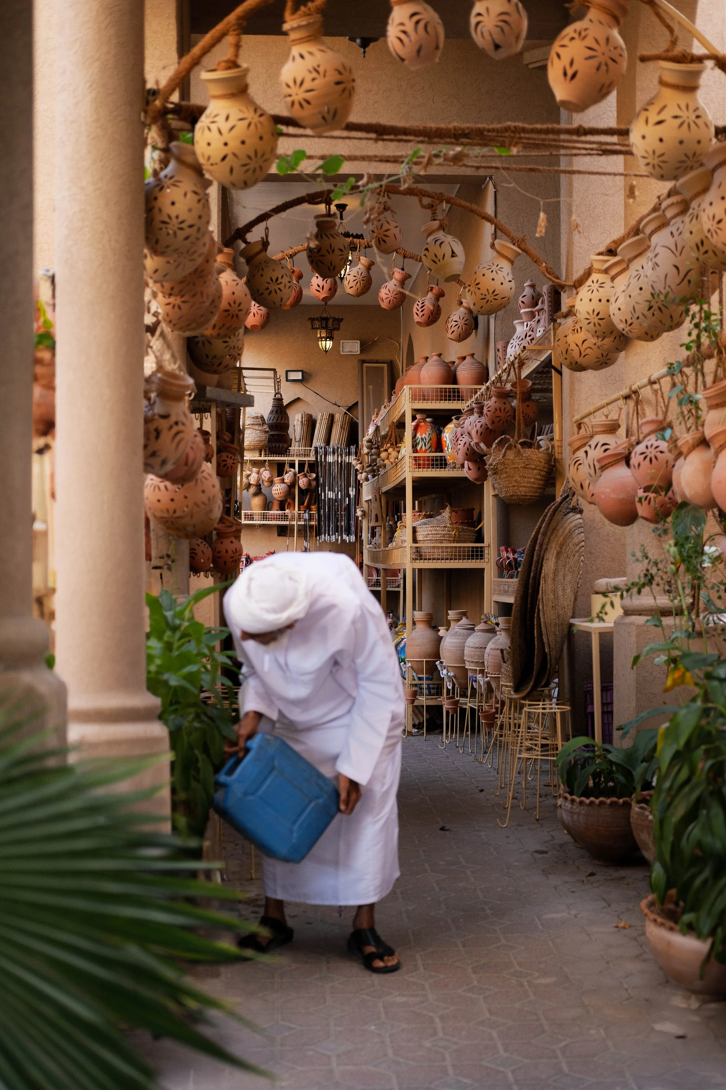 A person dressed in white traditional clothing watering plants in a shop that sells pottery, with clay jars and pots hanging from strings and arranged on shelves.