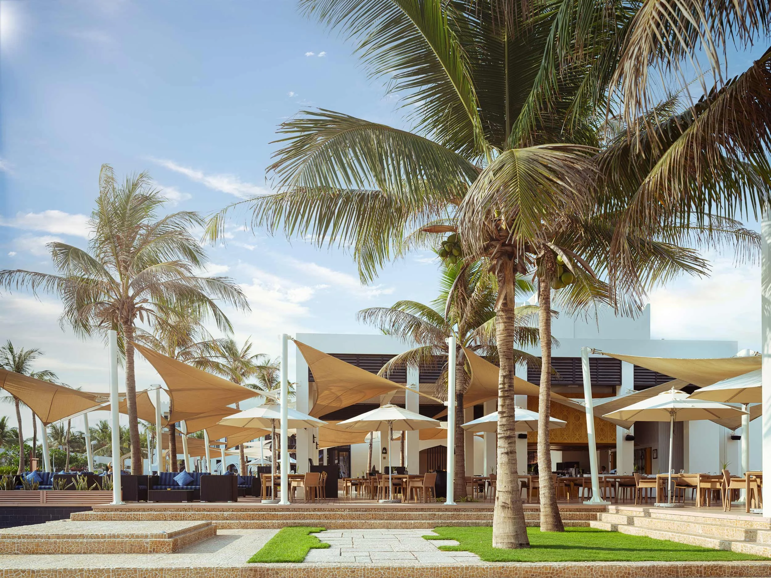 Outdoor seating area with tables, chairs, large umbrellas, and palm trees on a sunny day.