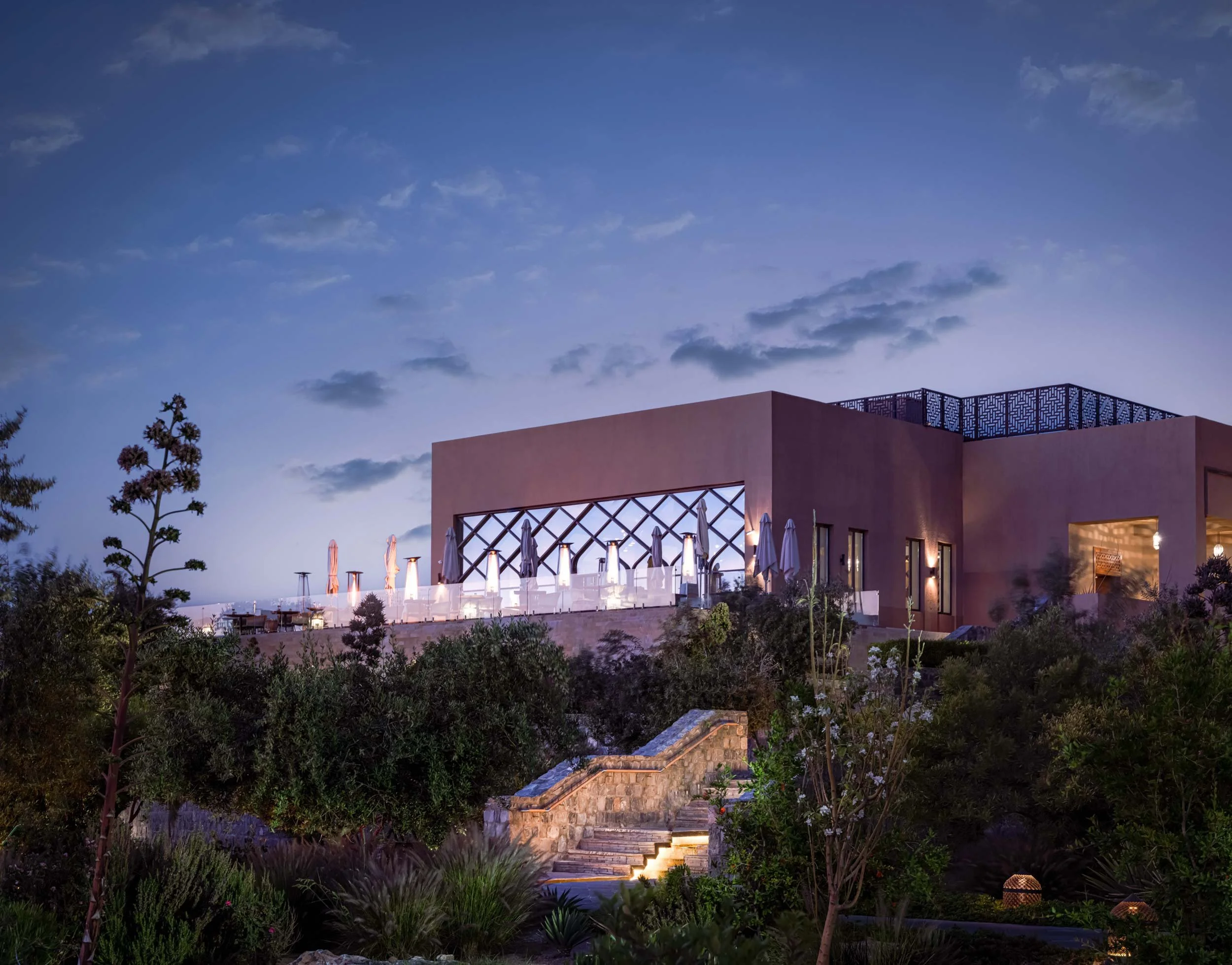 Modern building with large balcony and outdoor seating area, surrounded by greenery, under a blue sky at dusk
