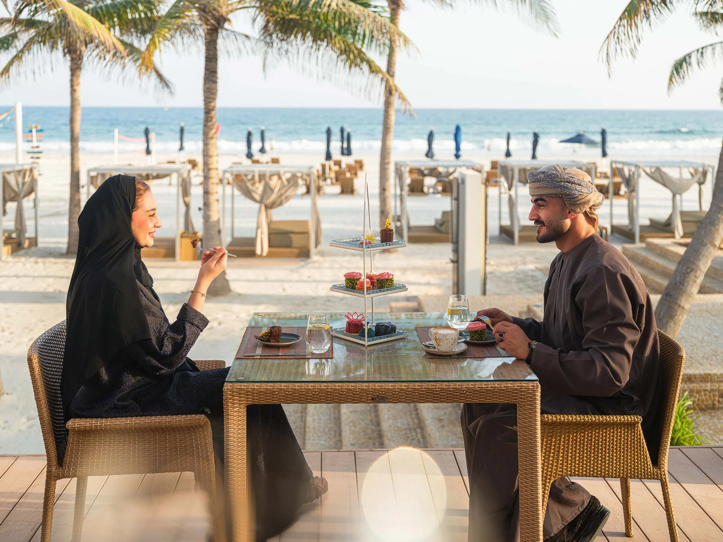 A man and woman dining at a seaside restaurant with ocean view in the background. They are enjoying desserts and smiling at each other, with palm trees and beach umbrellas behind them.