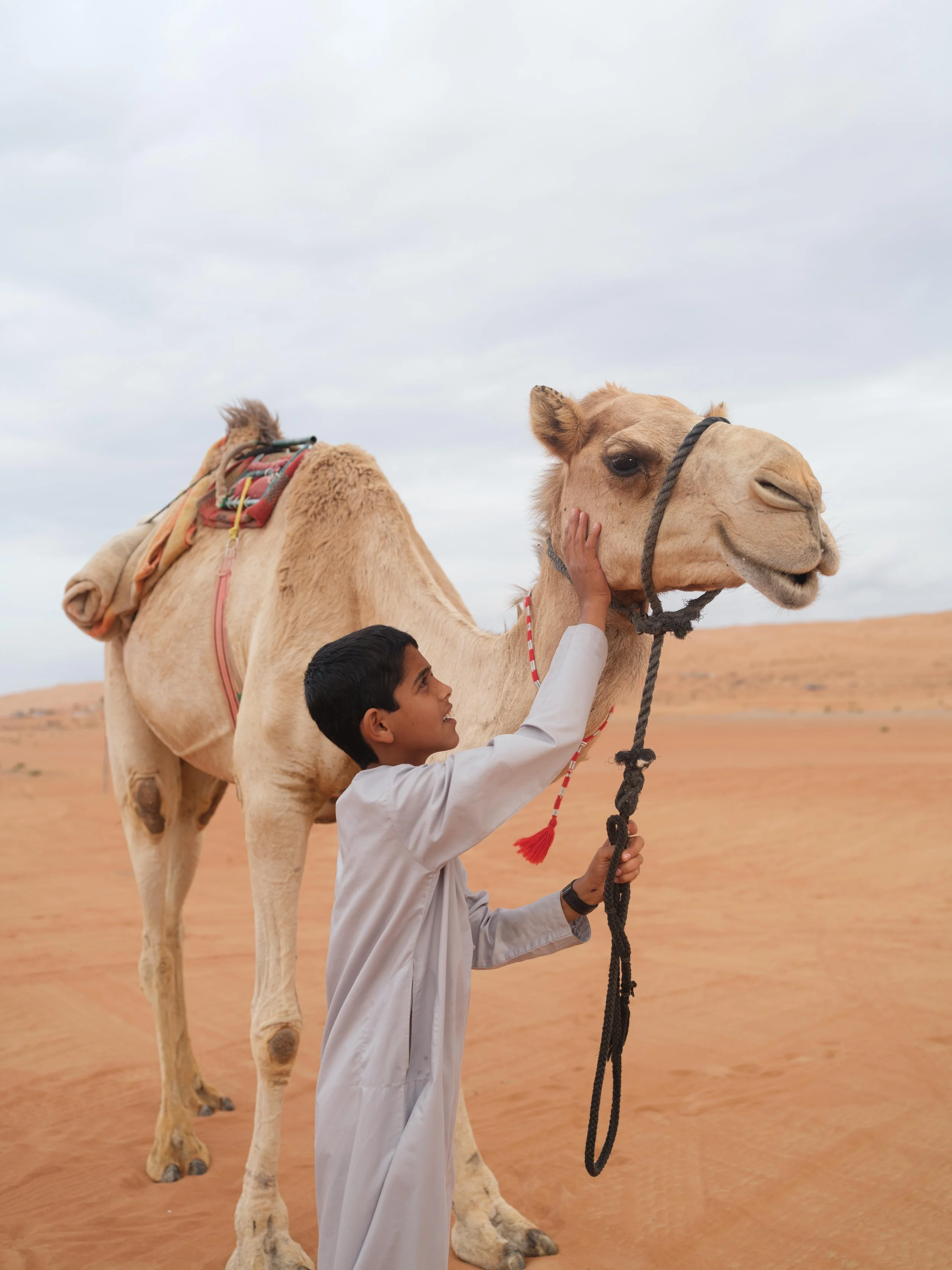 A boy in traditional clothing strokes a camel in the desert.