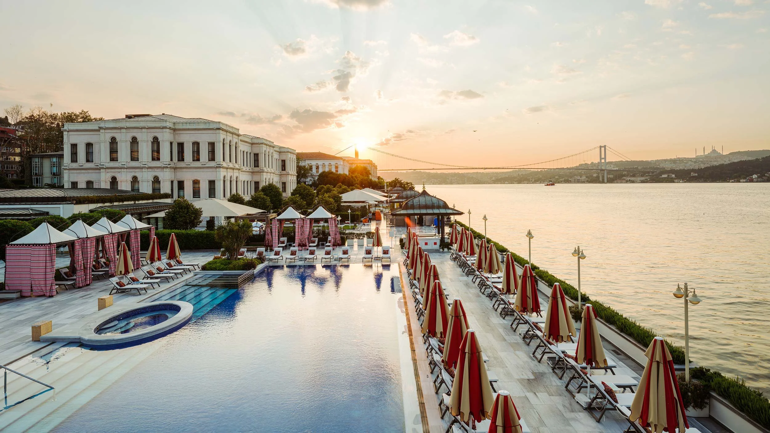 View of  luxurious poolside area of Four Seasons Bosphorus Hotel with red and beige umbrellas and lounge chairs by a river at sunset, with a bridge and hills in the background.