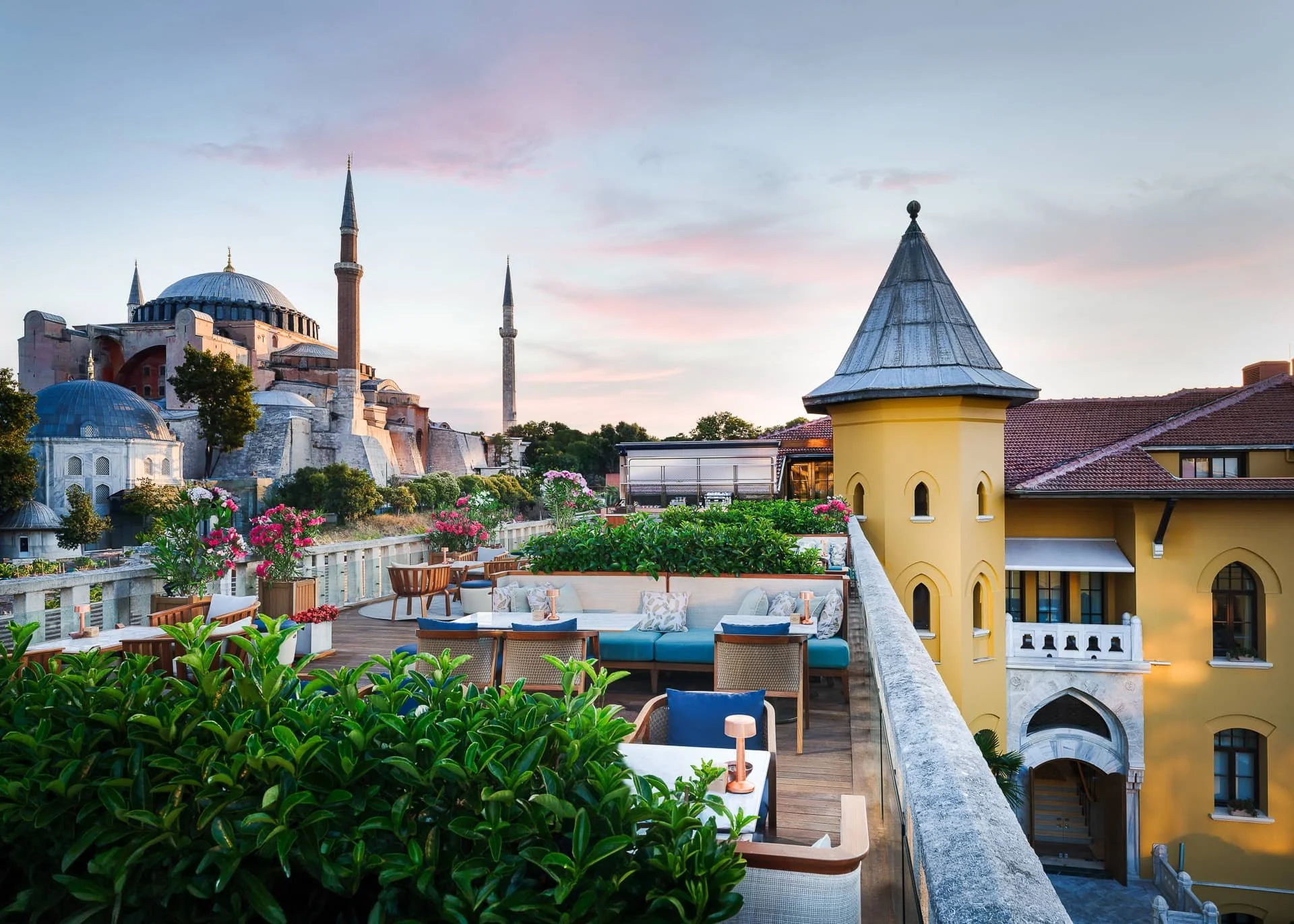 A rooftop terrace at Four Seasons Istanbul Hotel at Sultanahmet with seating, plants, and pink flowers, overlooking a Hagia Sophia with domes and minarets at sunset.