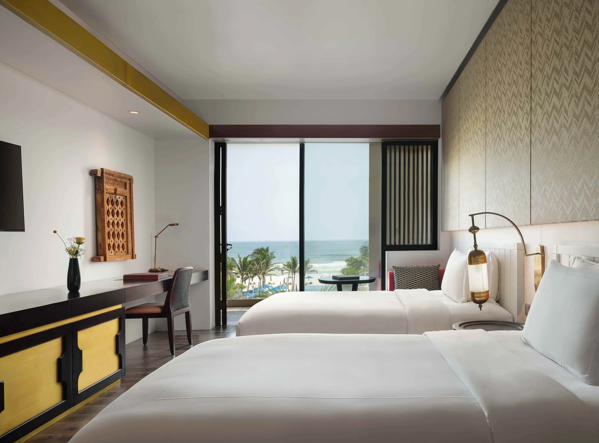 Modern hotel room with two beds, ocean view through sliding glass door, tropical trees outside, minimalist decor, and ambient lighting.