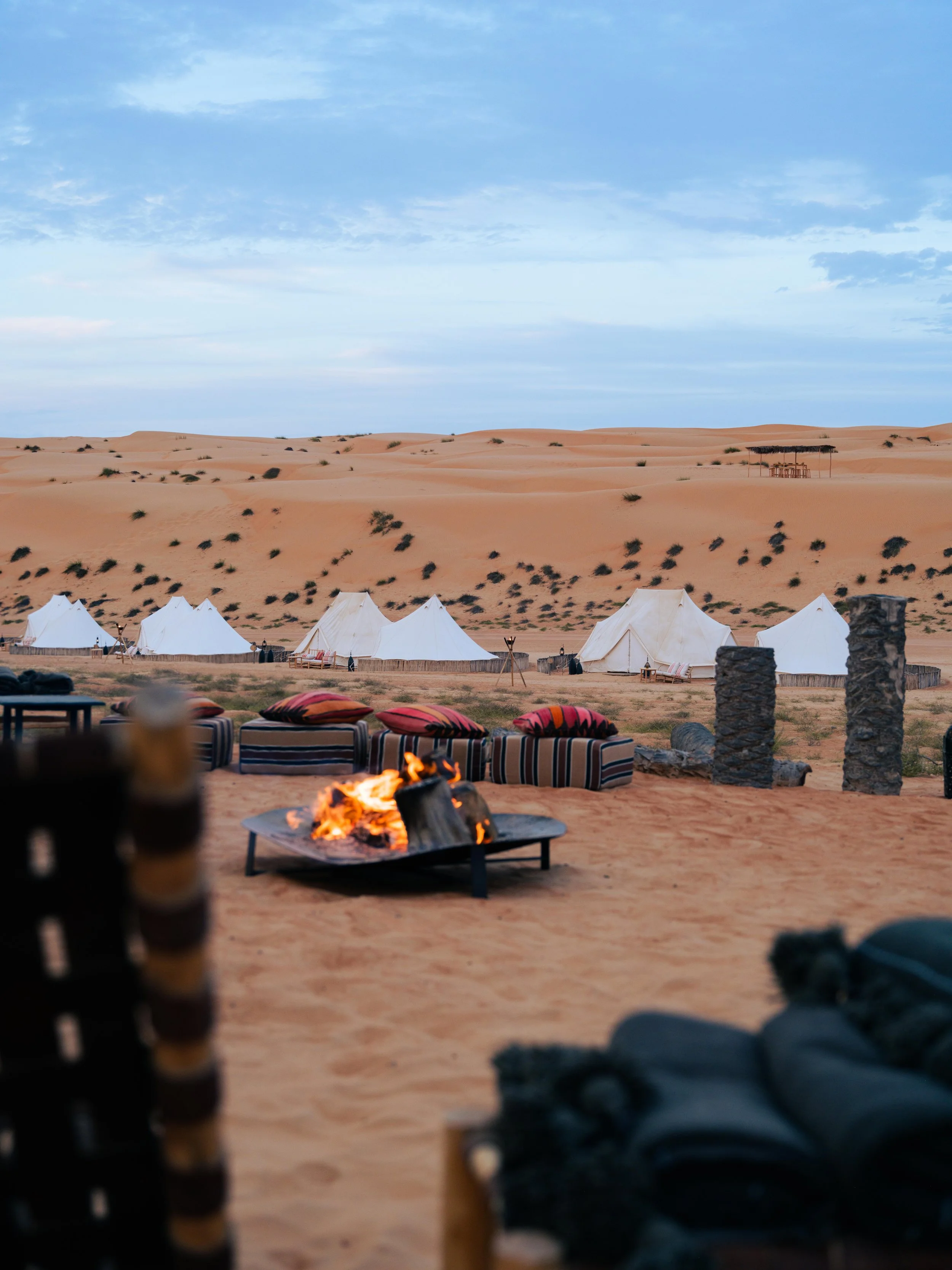 Desert camp with large tents, a burning campfire, and seating surrounded by sand dunes under a partly cloudy sky.