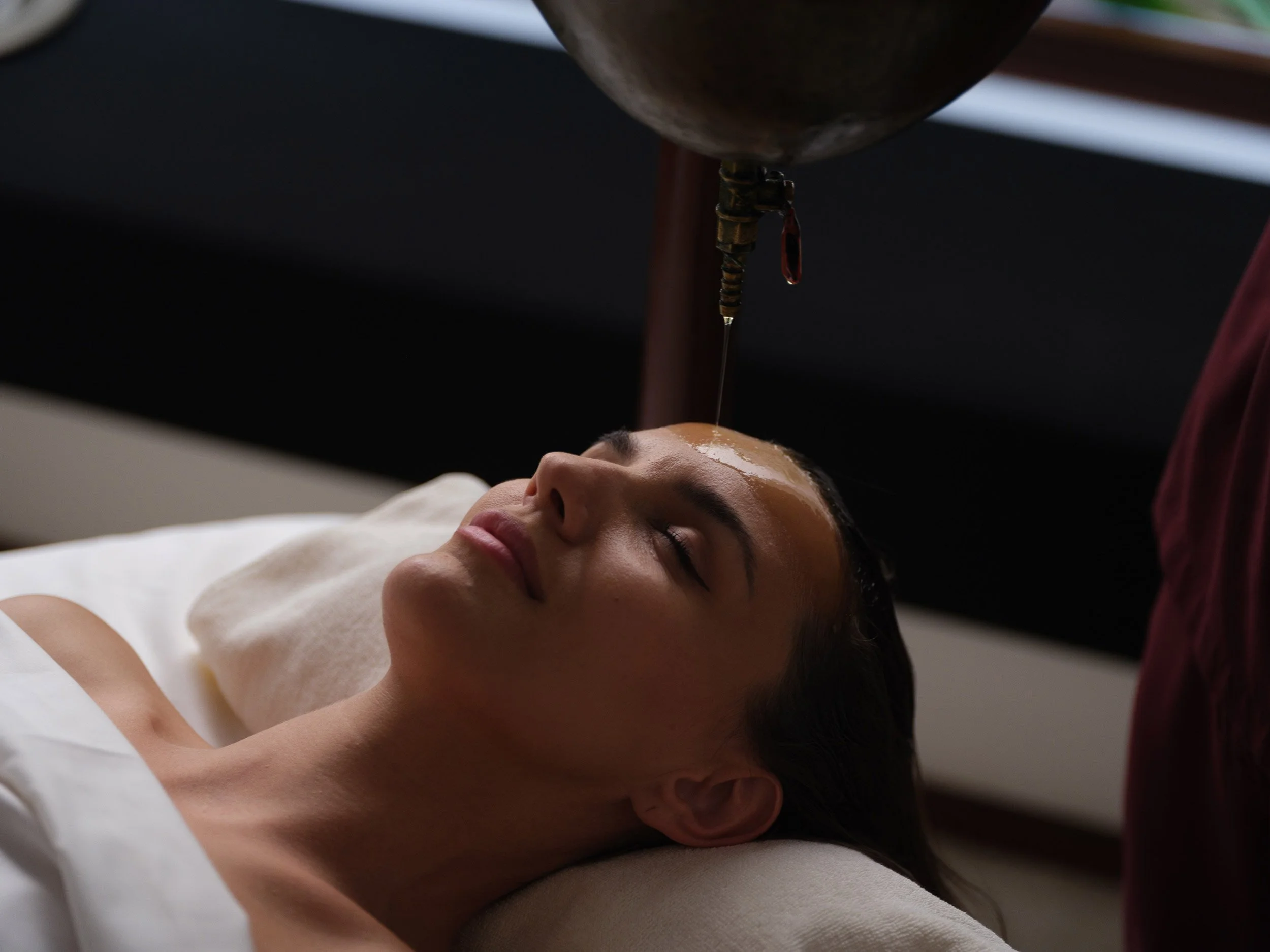 A woman lying down with eyes closed, receiving a facial treatment where a tool above her forehead is dripping a liquid onto her skin.