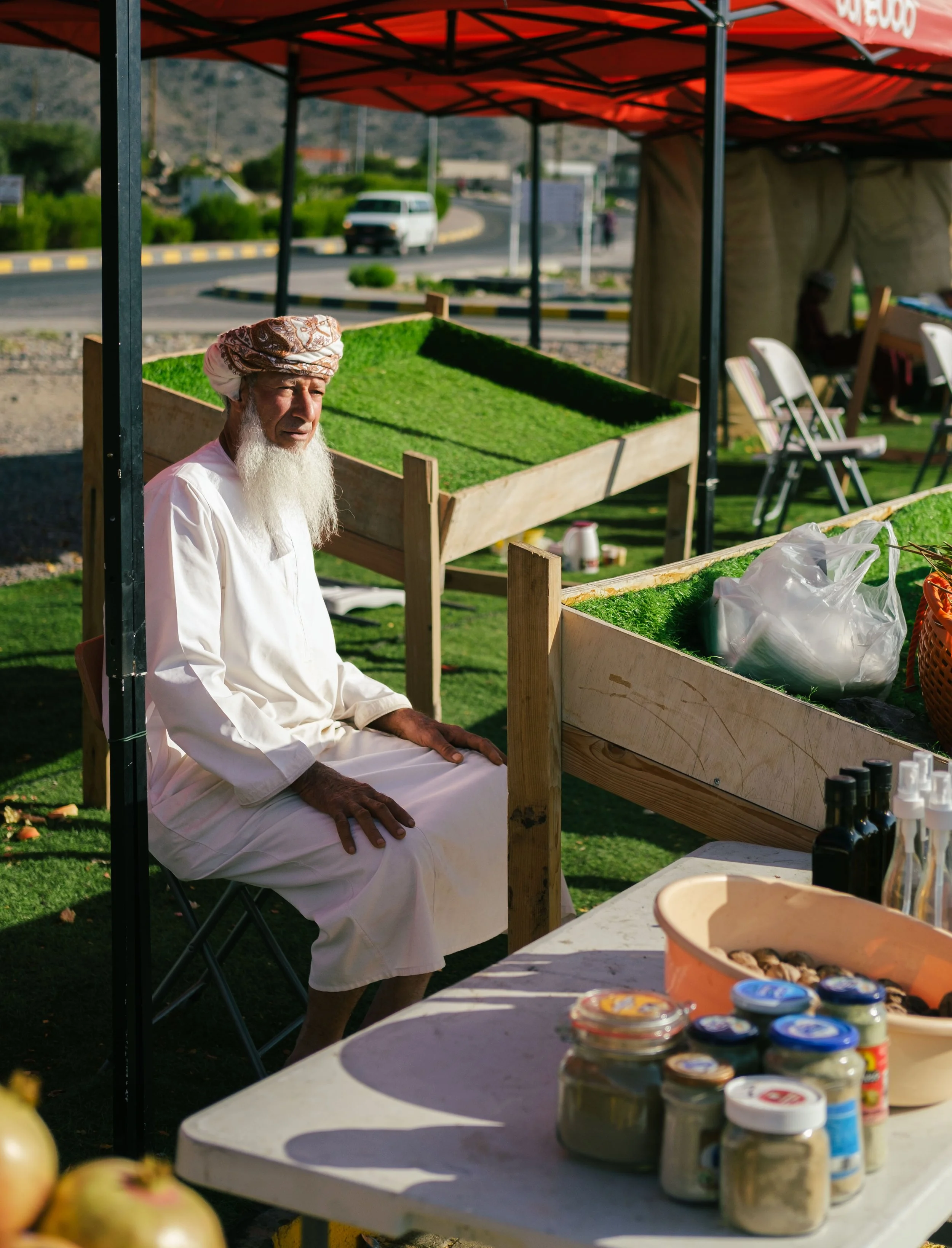 An elderly man with a white beard wearing traditional attire and a patterned turban, sitting under a red canopy at a market stall with jars of goods and apples on a table.