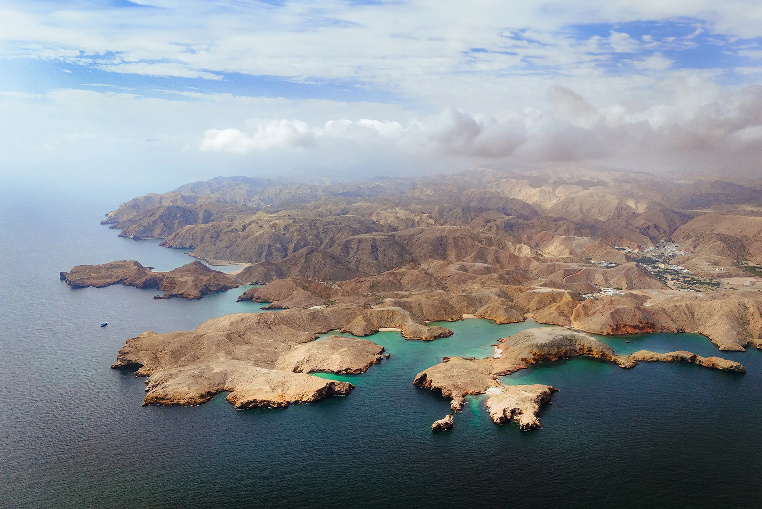 Aerial view of a rugged coastline with small islands and bays, surrounded by arid, mountainous terrain under a partly cloudy sky.