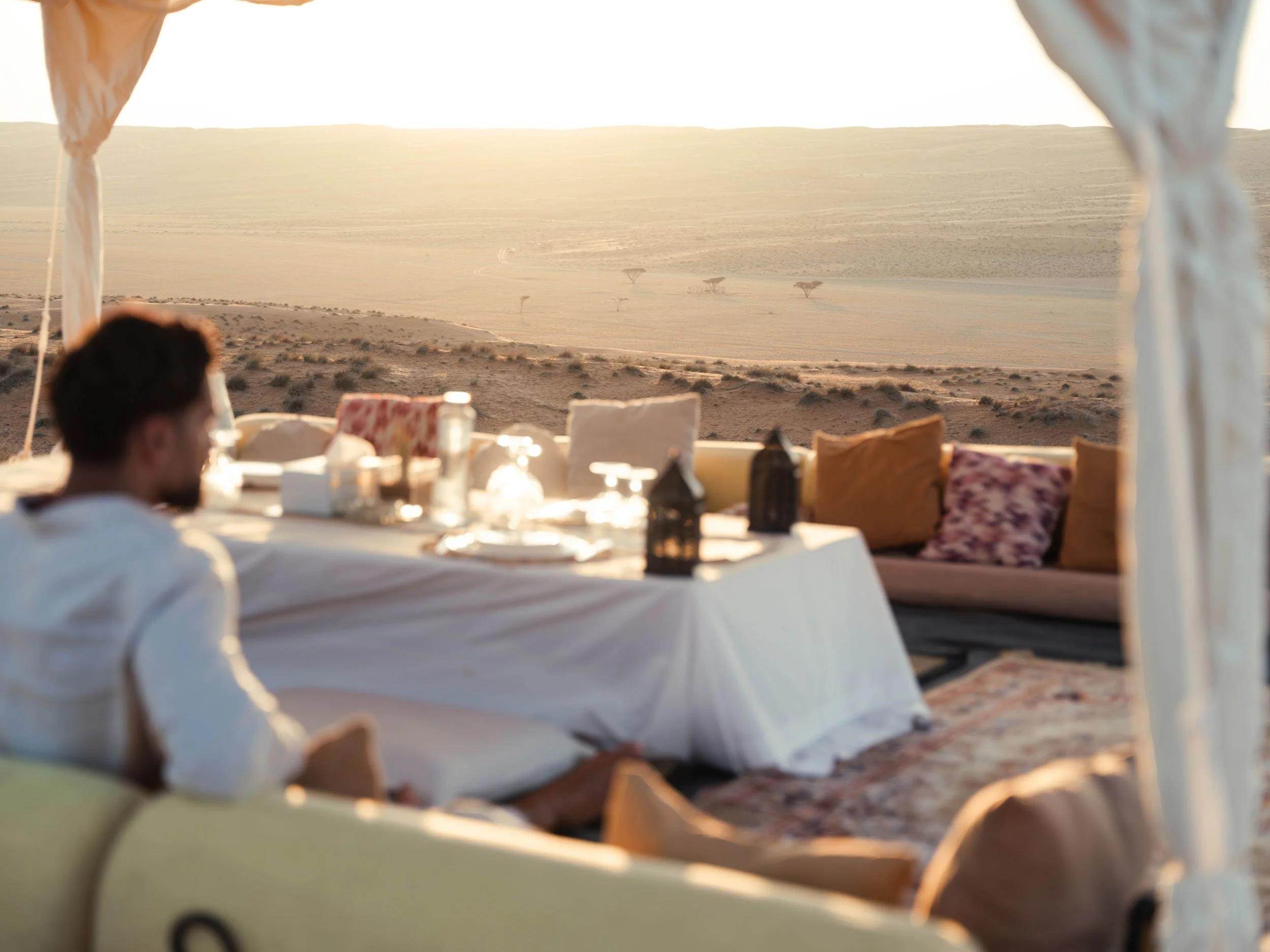 A person sitting at a table inside a white tent in a desert, with a view of desert of Wahiba sands during sunset.
