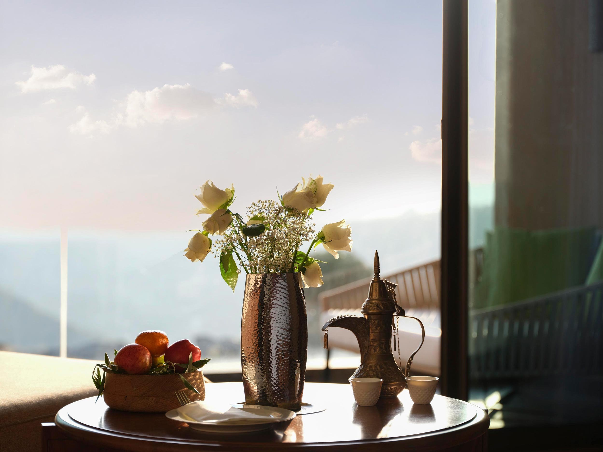 A round wooden table with a bowl of apples, a tall vase of white flowers, and a metal pitcher with two cups, illuminated by natural light in a room with a large window and a scenic view outside.