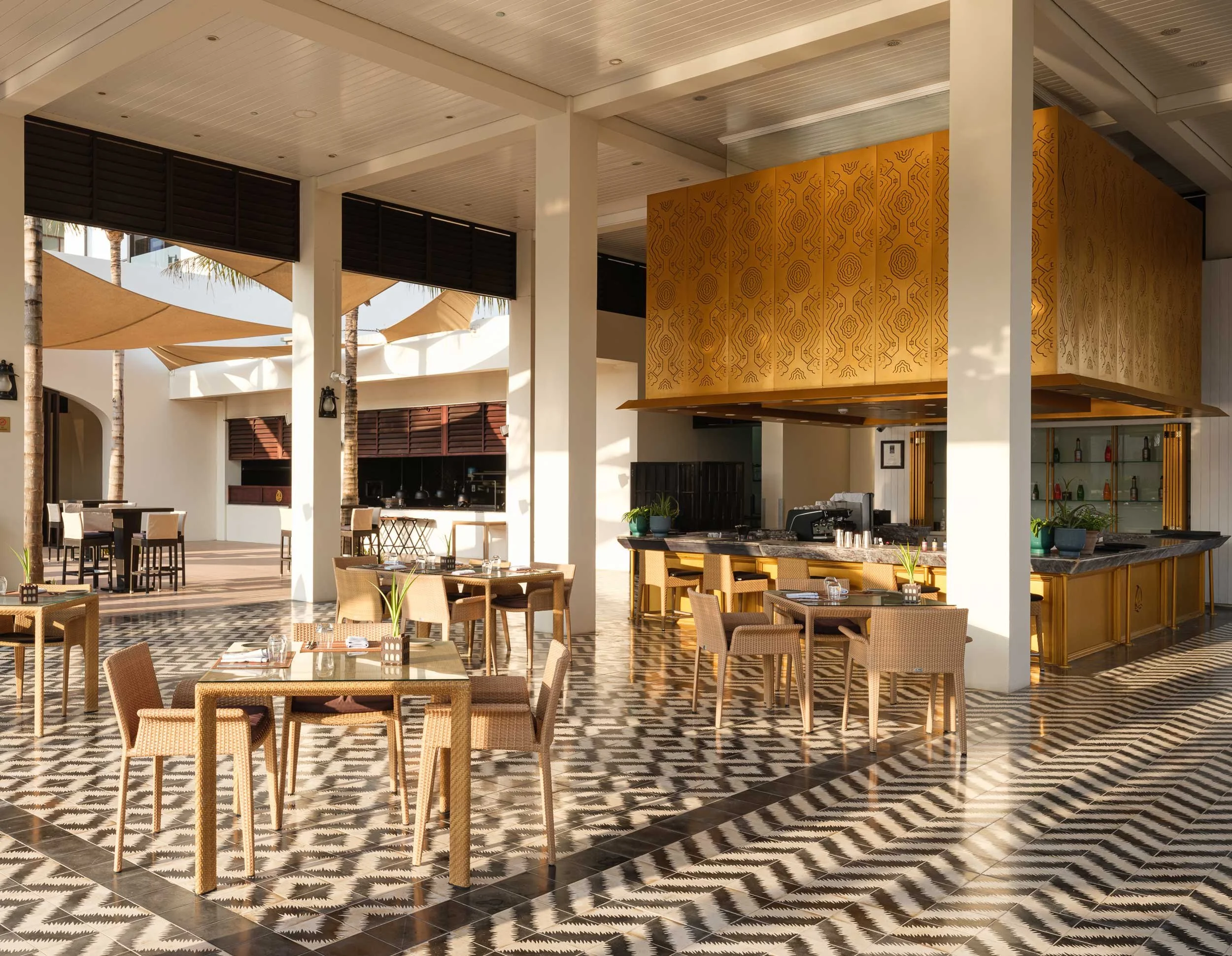 An outdoor restaurant patio with tables, chairs, and a bar area under shade sails, featuring patterned tile flooring and tropical palm trees.