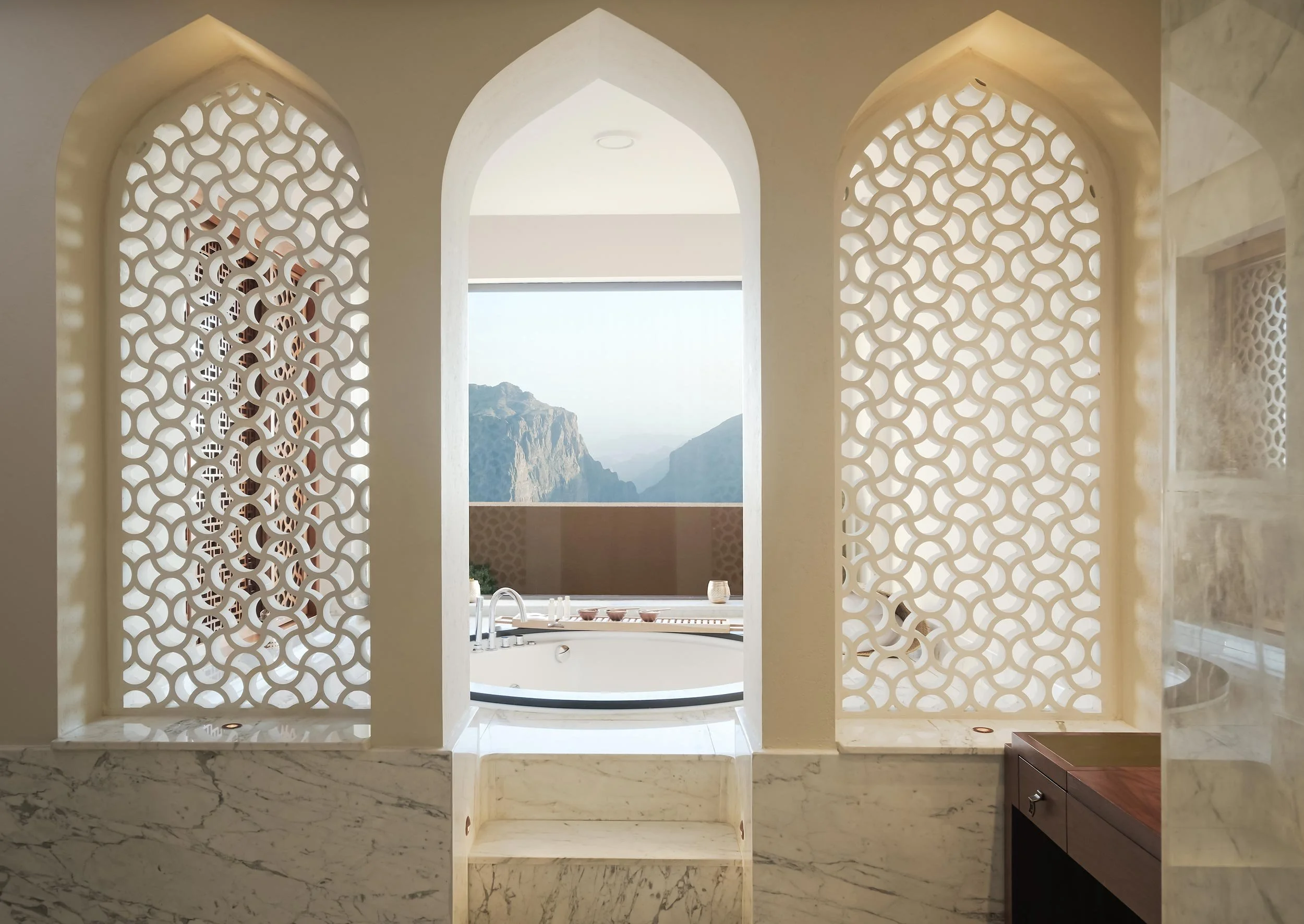 View of a luxurious bathroom with geometric lattice screens, a marble ledge, and a whirlpool tub overlooking mountainous landscape through a large window.