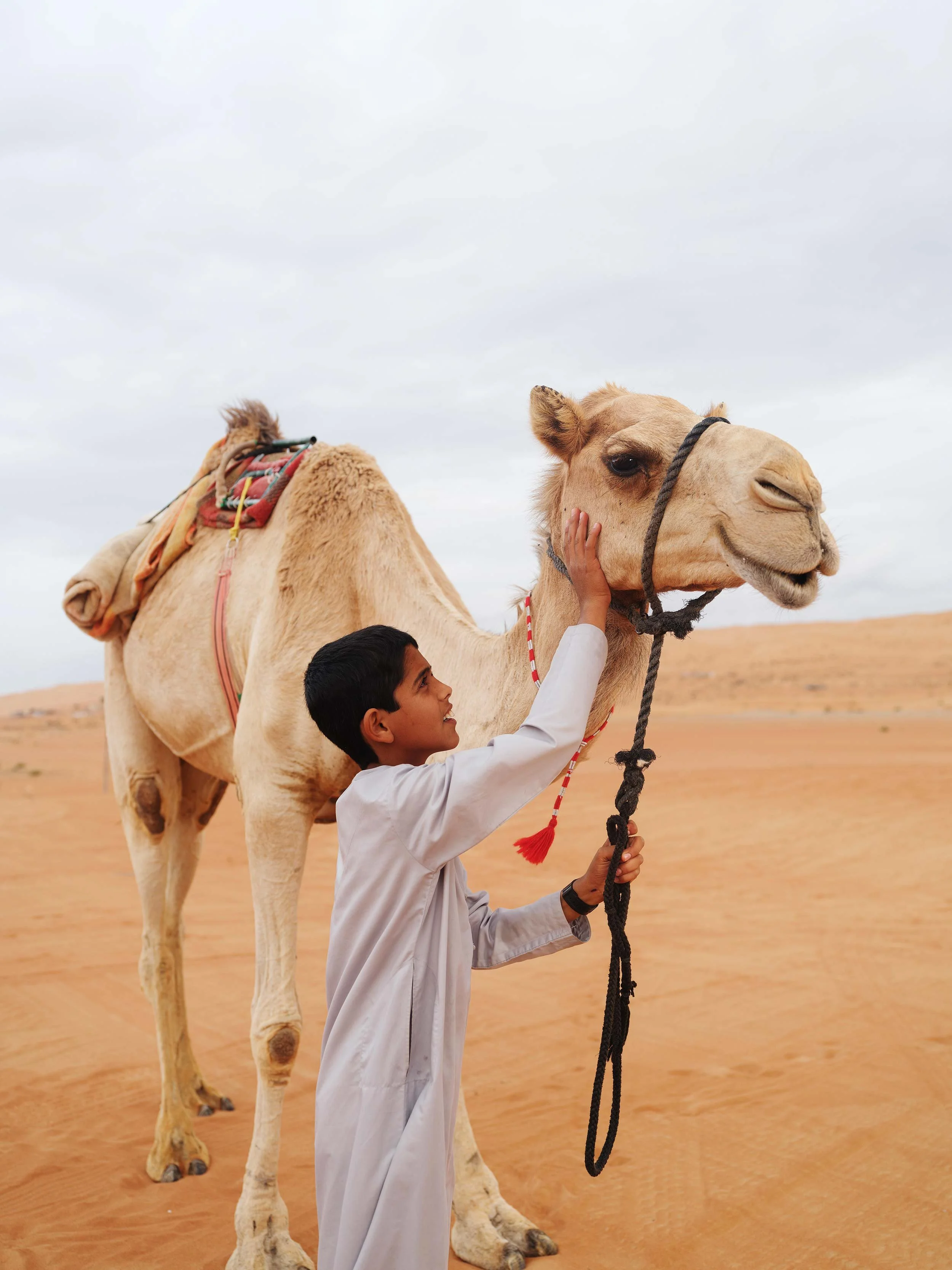 A young boy in traditional white clothing petting a camel in a desert landscape under a cloudy sky.